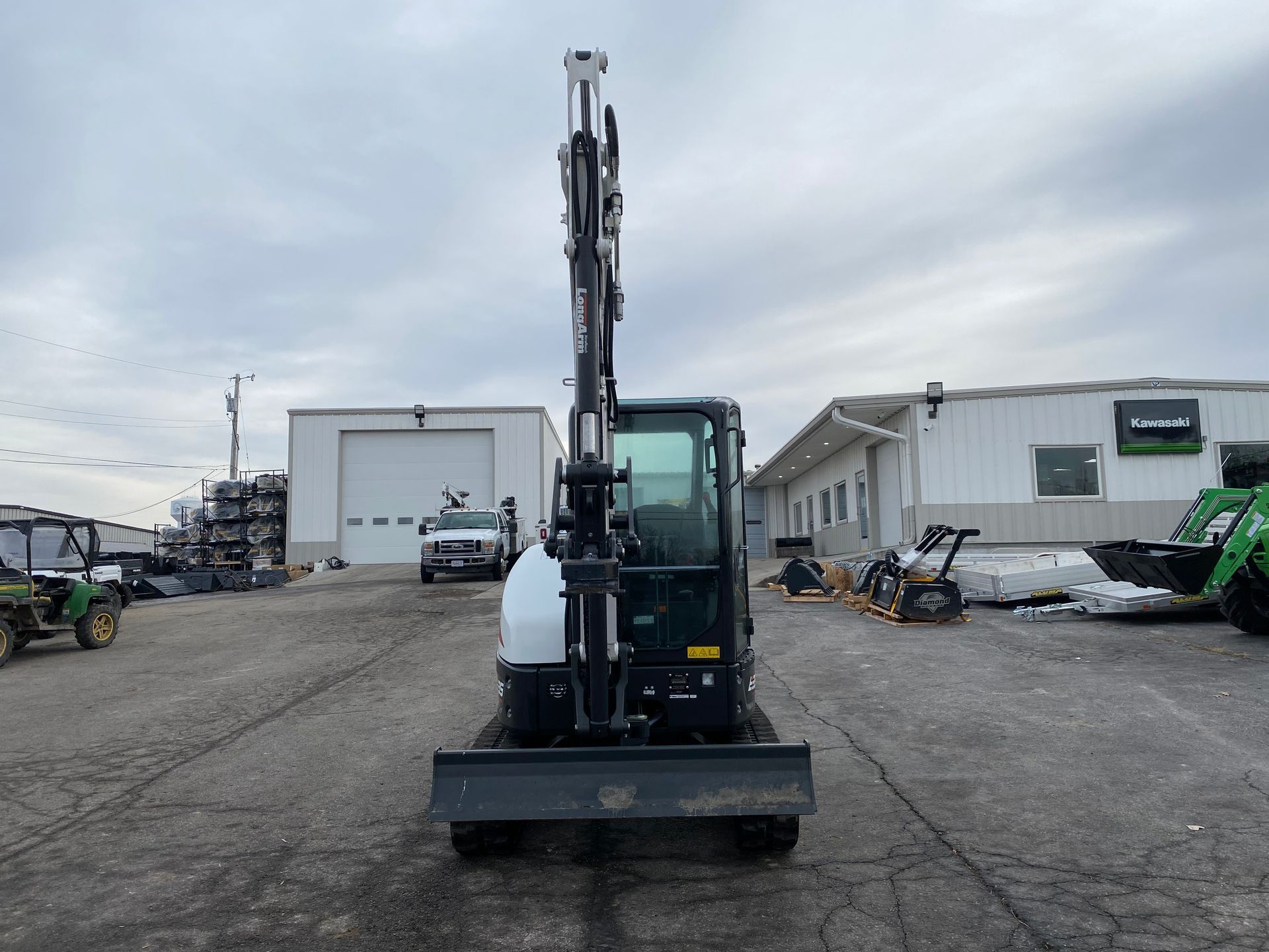 Mini excavator with arm raised against a cloudy sky, parked outside buildings.