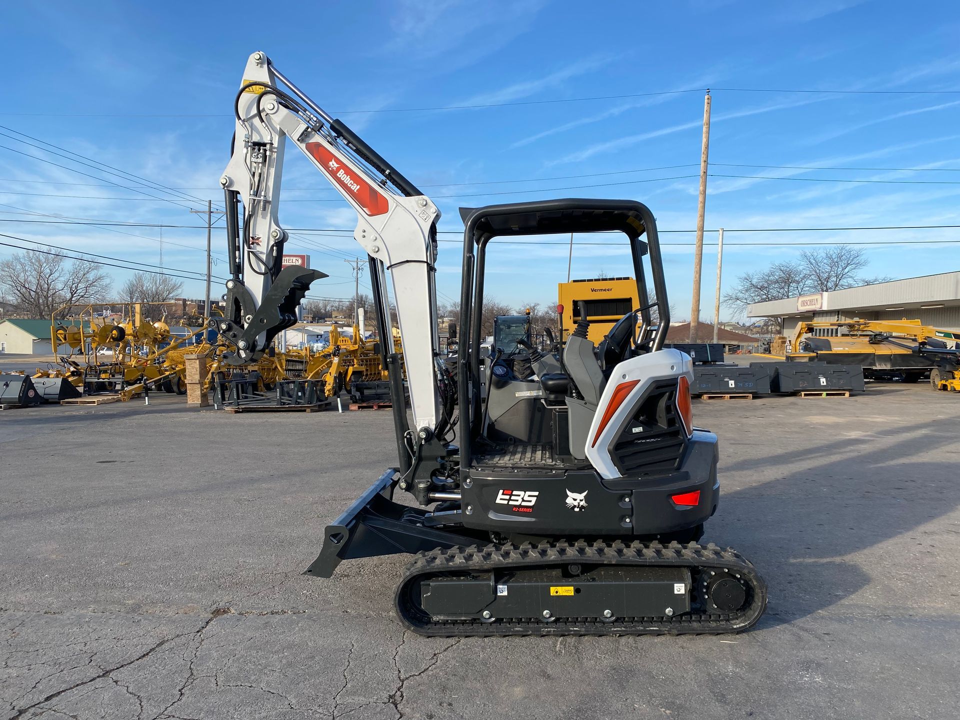 Bobcat mini excavator on pavement, arm raised, gray and white with orange accents, sunny outdoor setting.