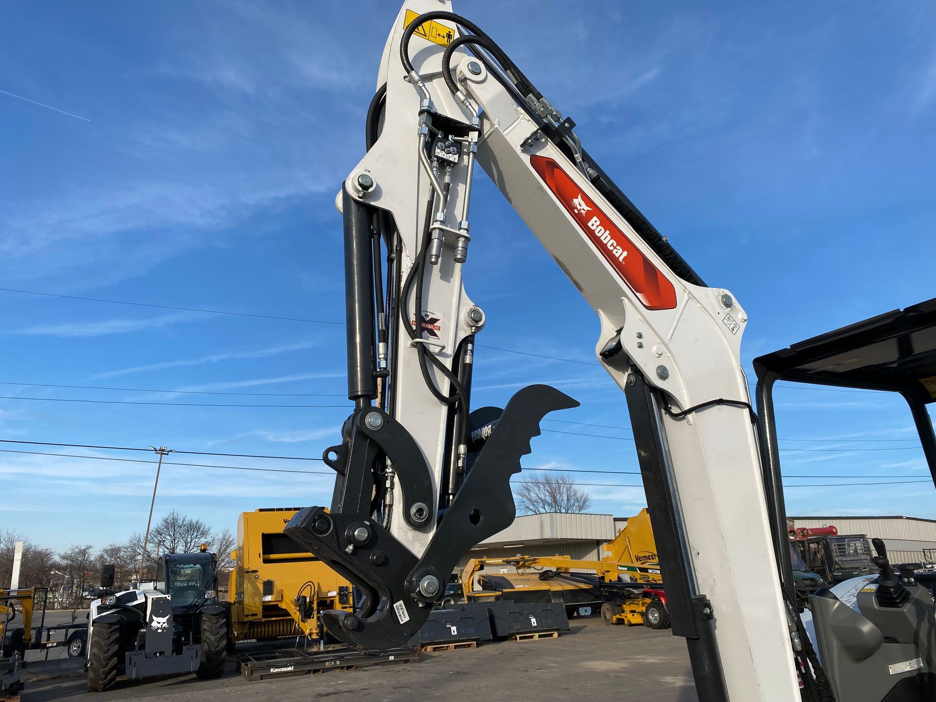 White Bobcat grapple attachment raised against a blue sky, in a machinery yard.