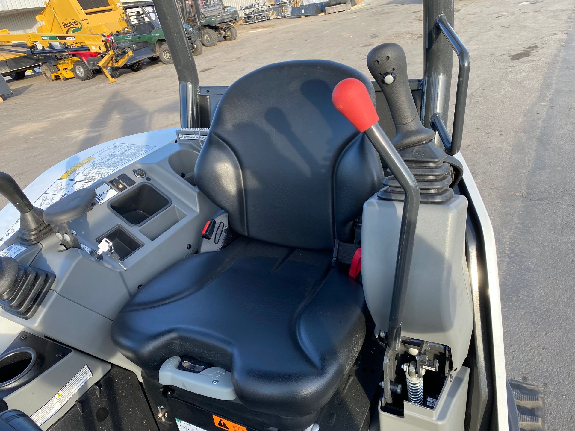 Interior view of a construction machine's seat and controls. Black seat, red joystick, grey dashboard.