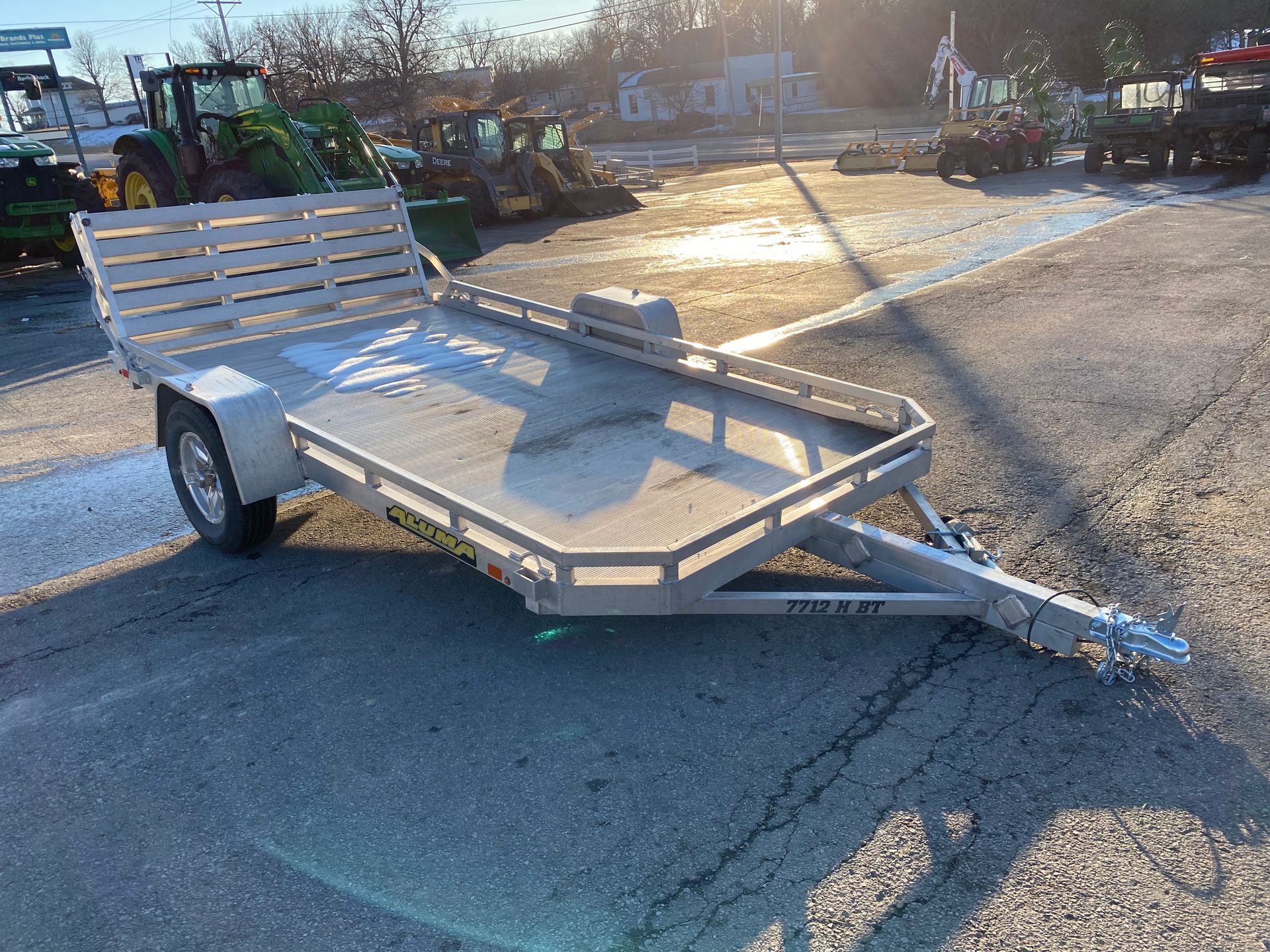 Silver utility trailer on asphalt in an outdoor lot, with equipment in the background.