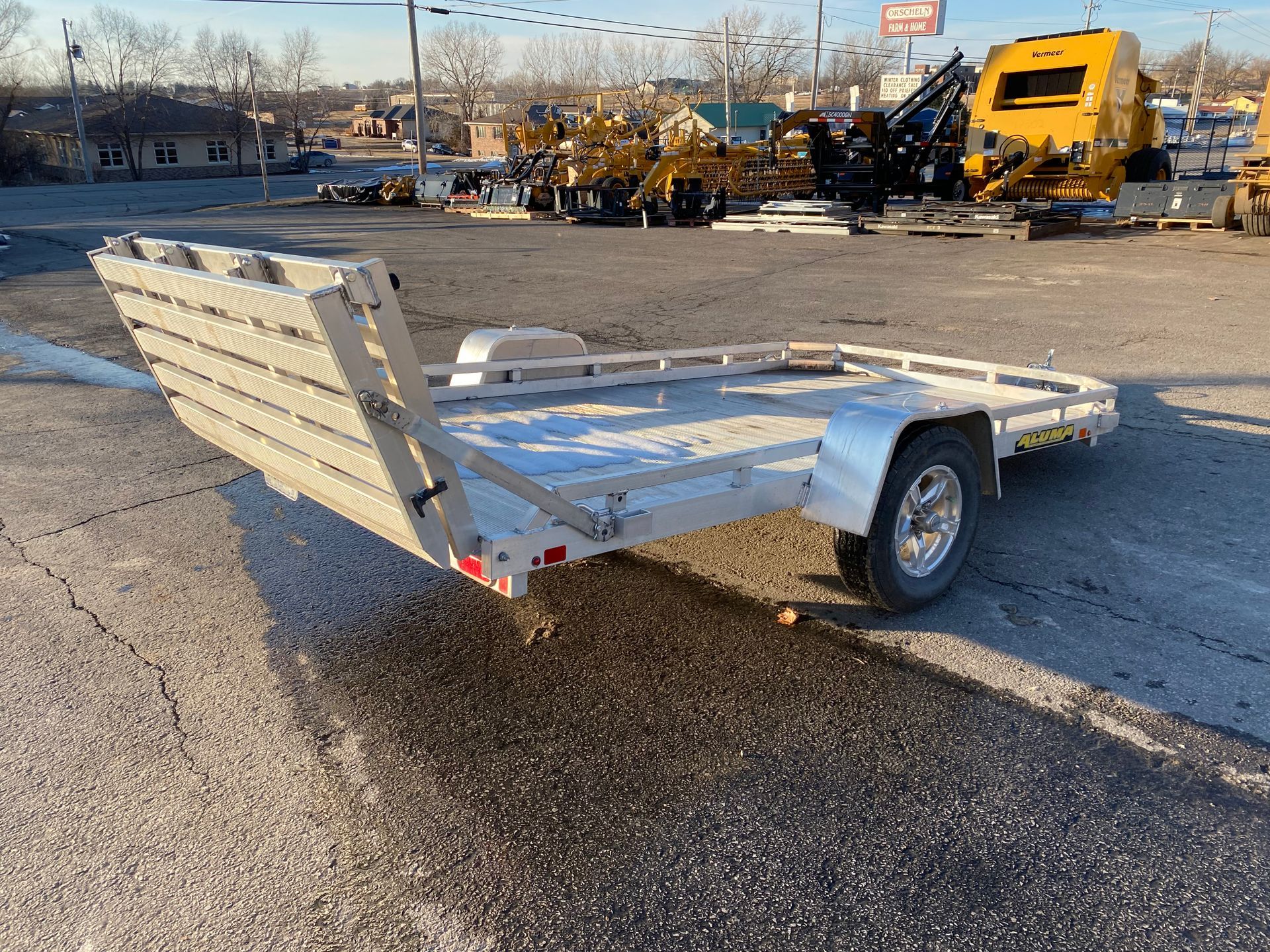 A silver utility trailer with a ramp, parked on an asphalt lot. Other equipment is in the background.