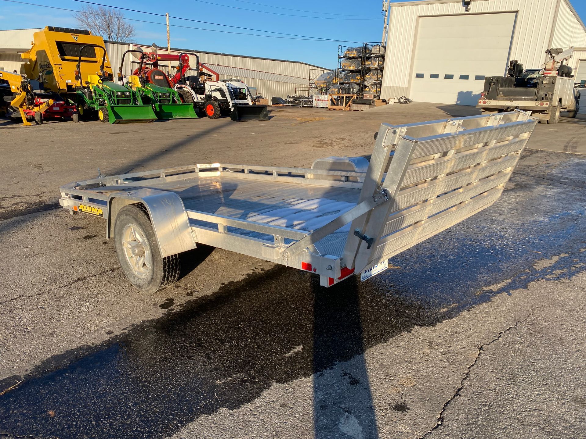 Silver utility trailer with ramp, parked outside a building, near other equipment.