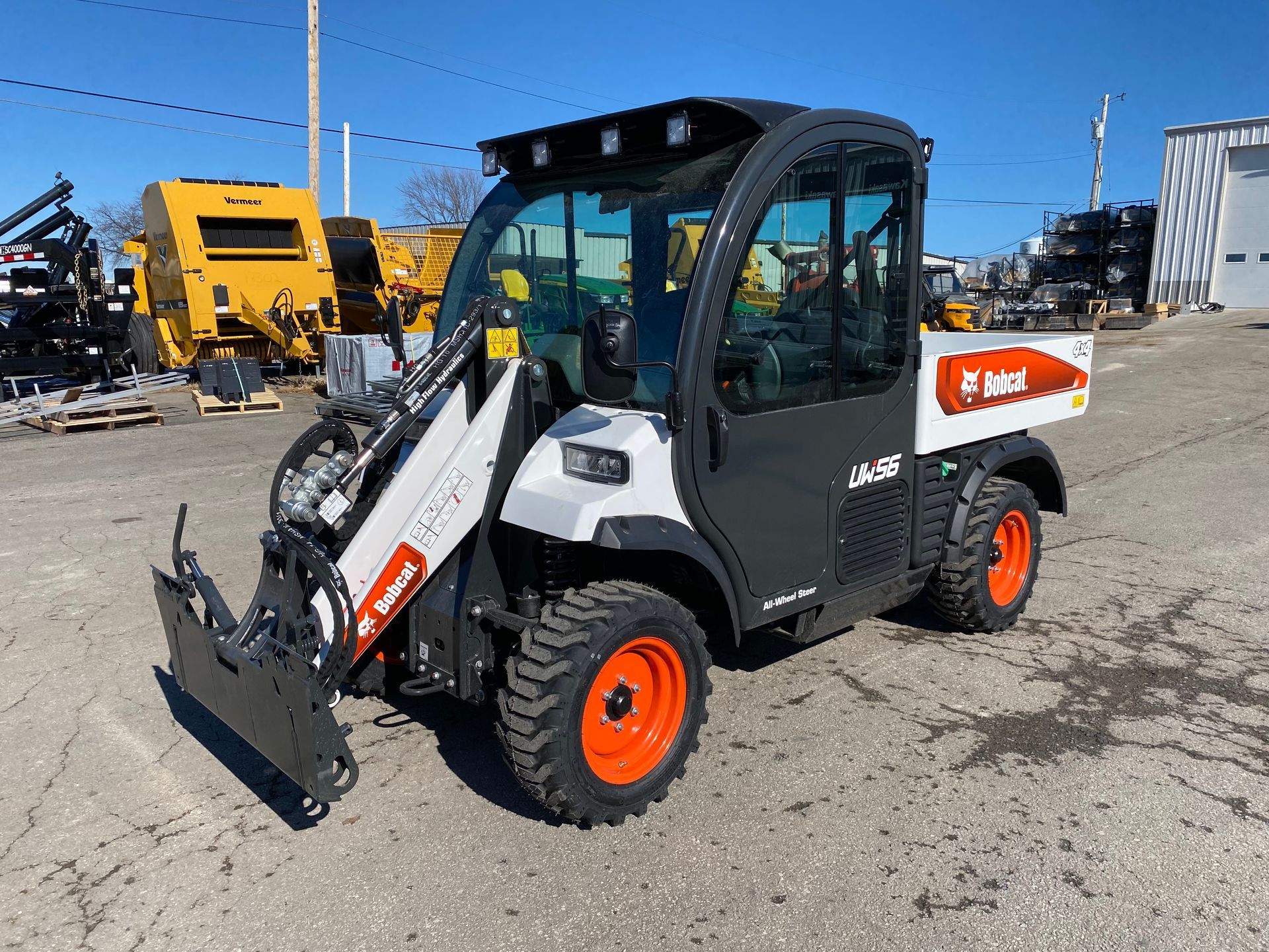 Bobcat utility vehicle with loader and enclosed cab on pavement. Gray, black, and white colors, orange wheels.