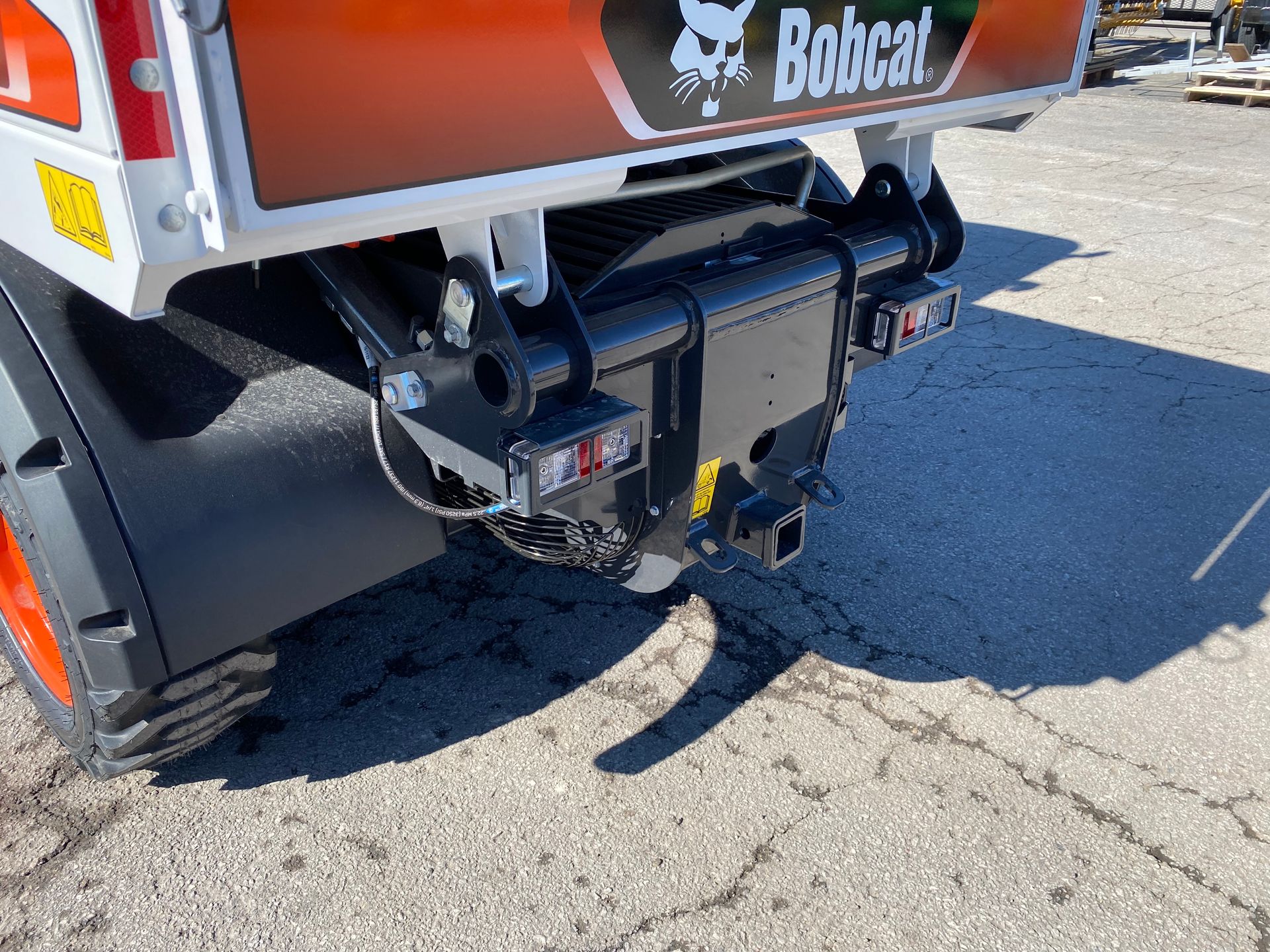 Rear view of a Bobcat attachment. Features a hitch, lights, and a white, black, and orange color scheme.