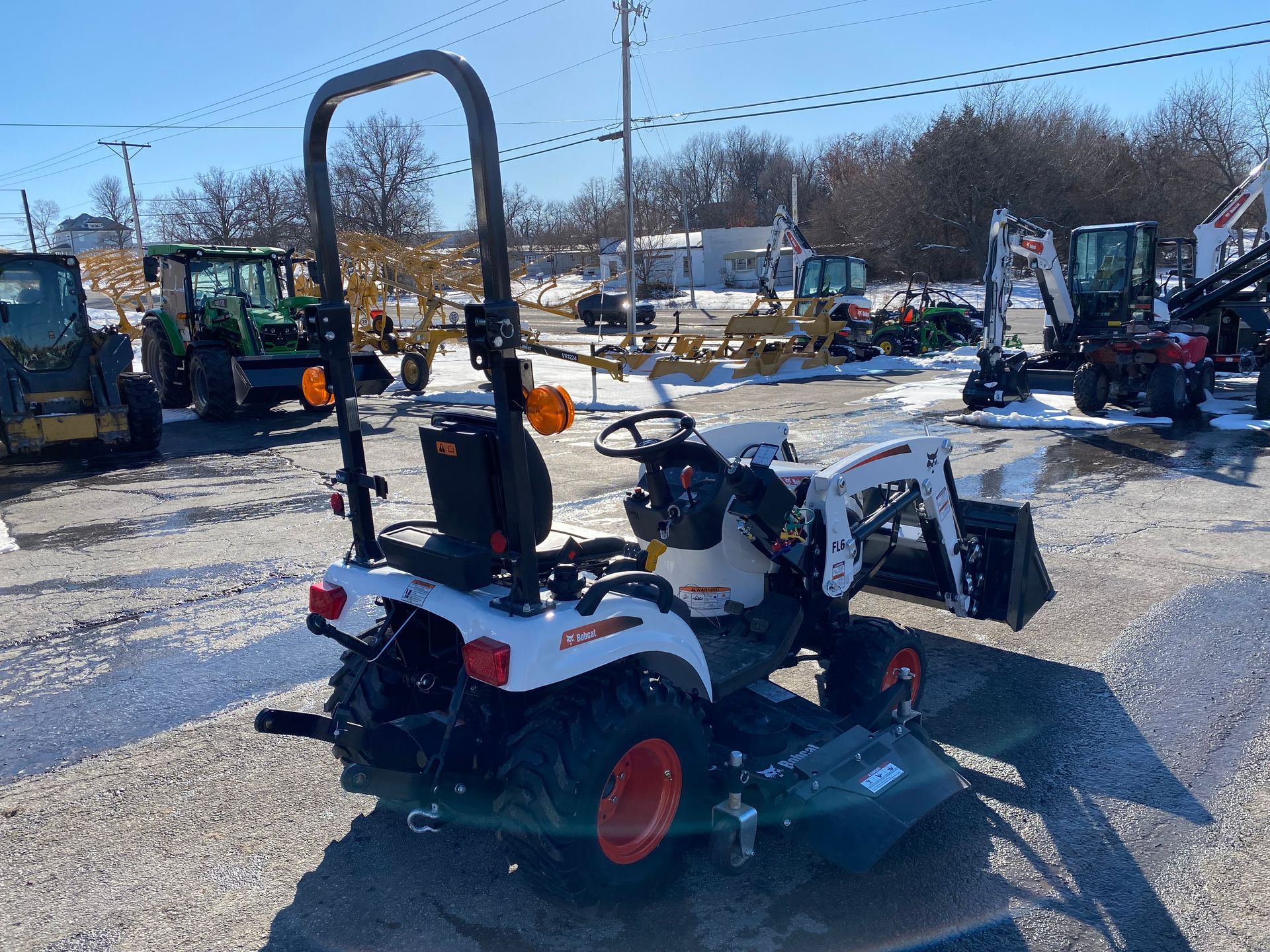 White Bobcat tractor with orange wheels, parked outdoors on a sunny day with snow and other machinery.