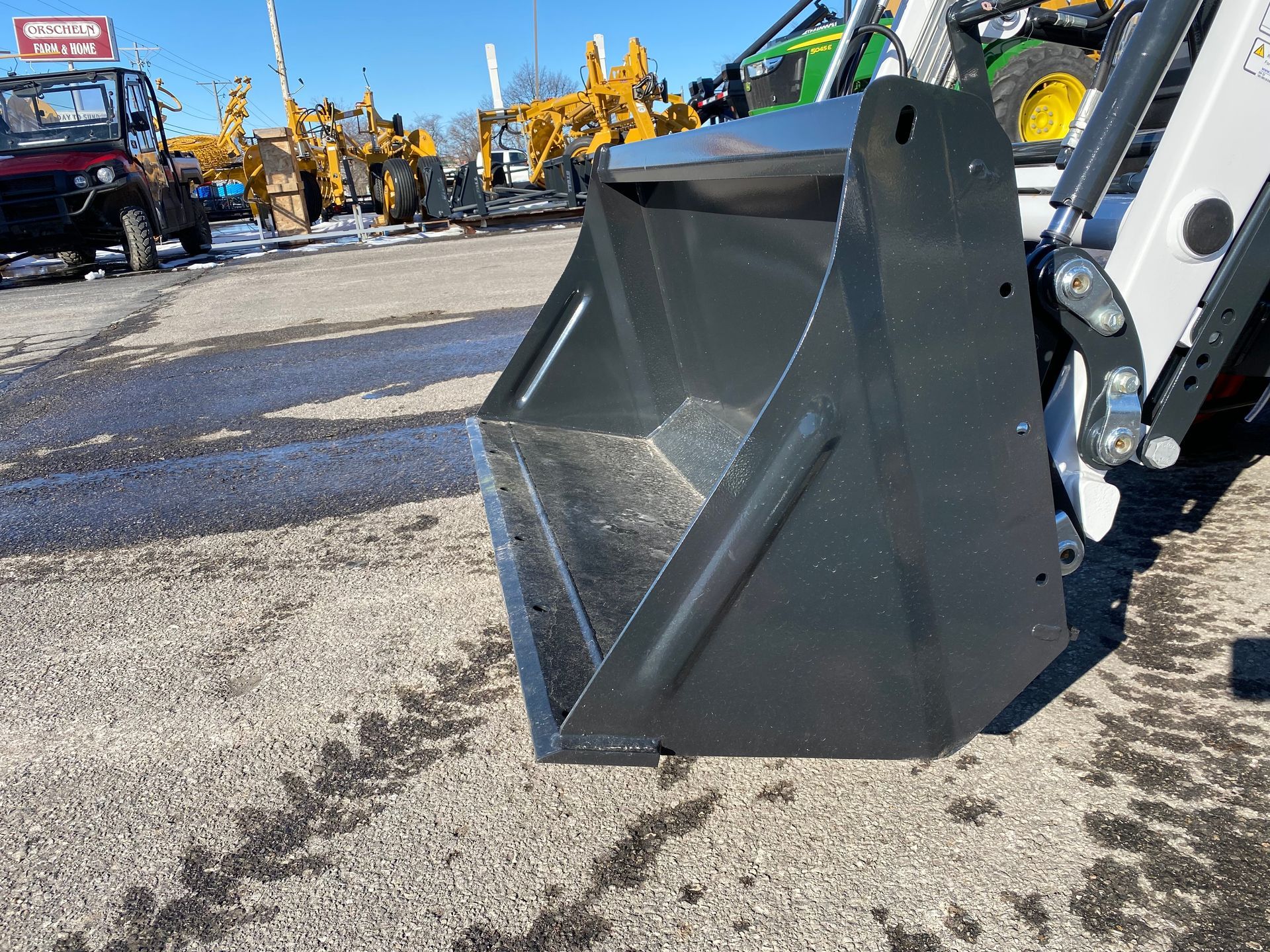 Black bucket attachment on a small tractor, outside on an asphalt lot.