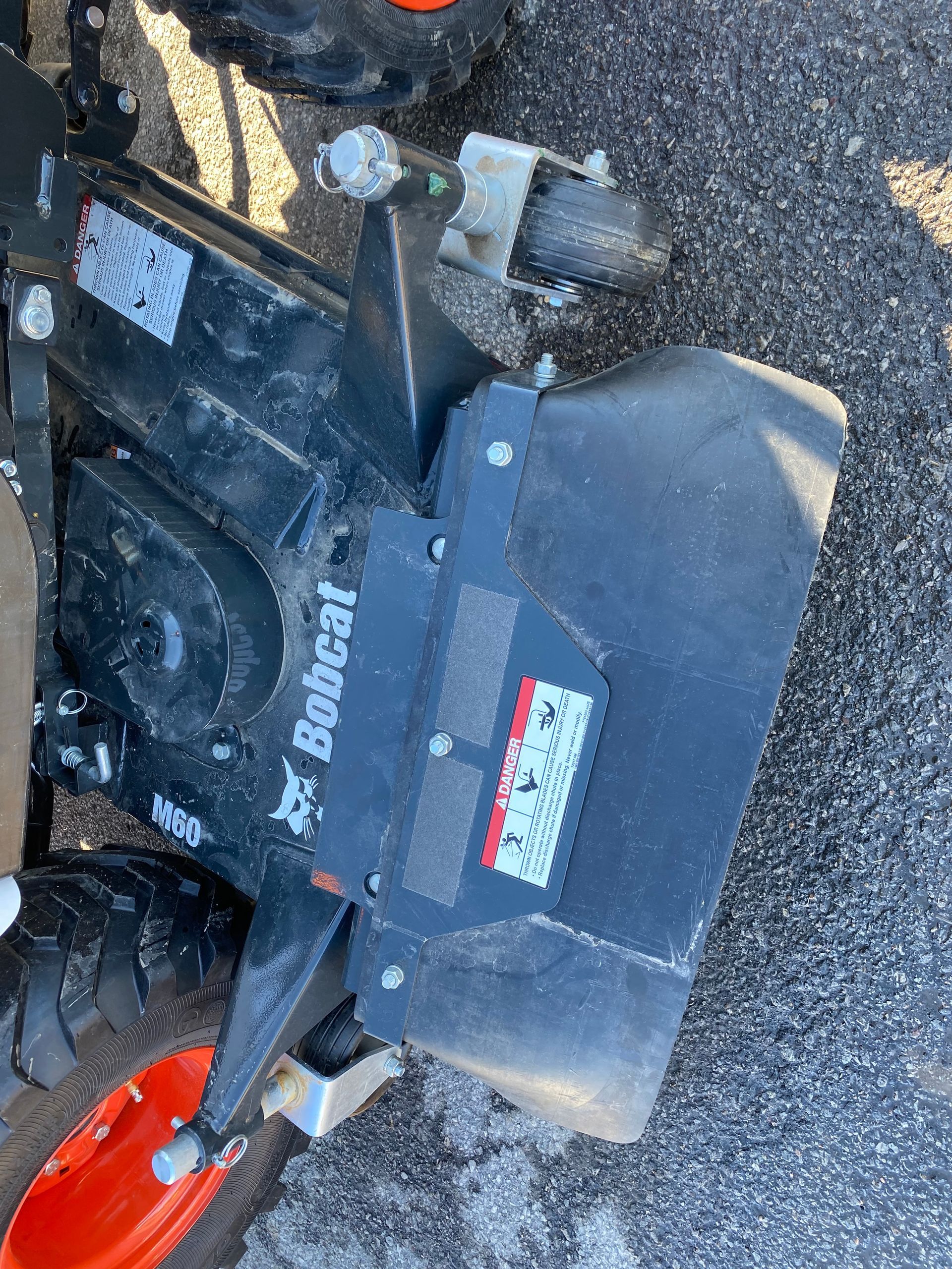Bobcat lawn mower attachment, black and silver, attached to a vehicle's wheel, with a safety warning label.