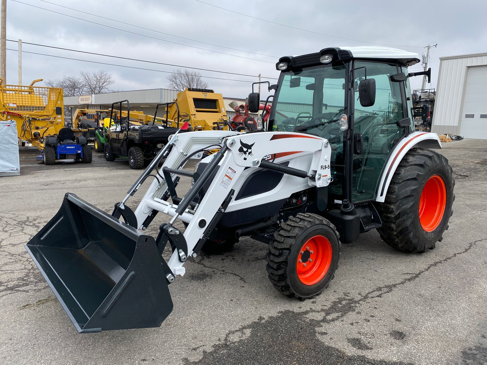 White and black Bobcat tractor with a front-end loader on a paved surface, other equipment in background.