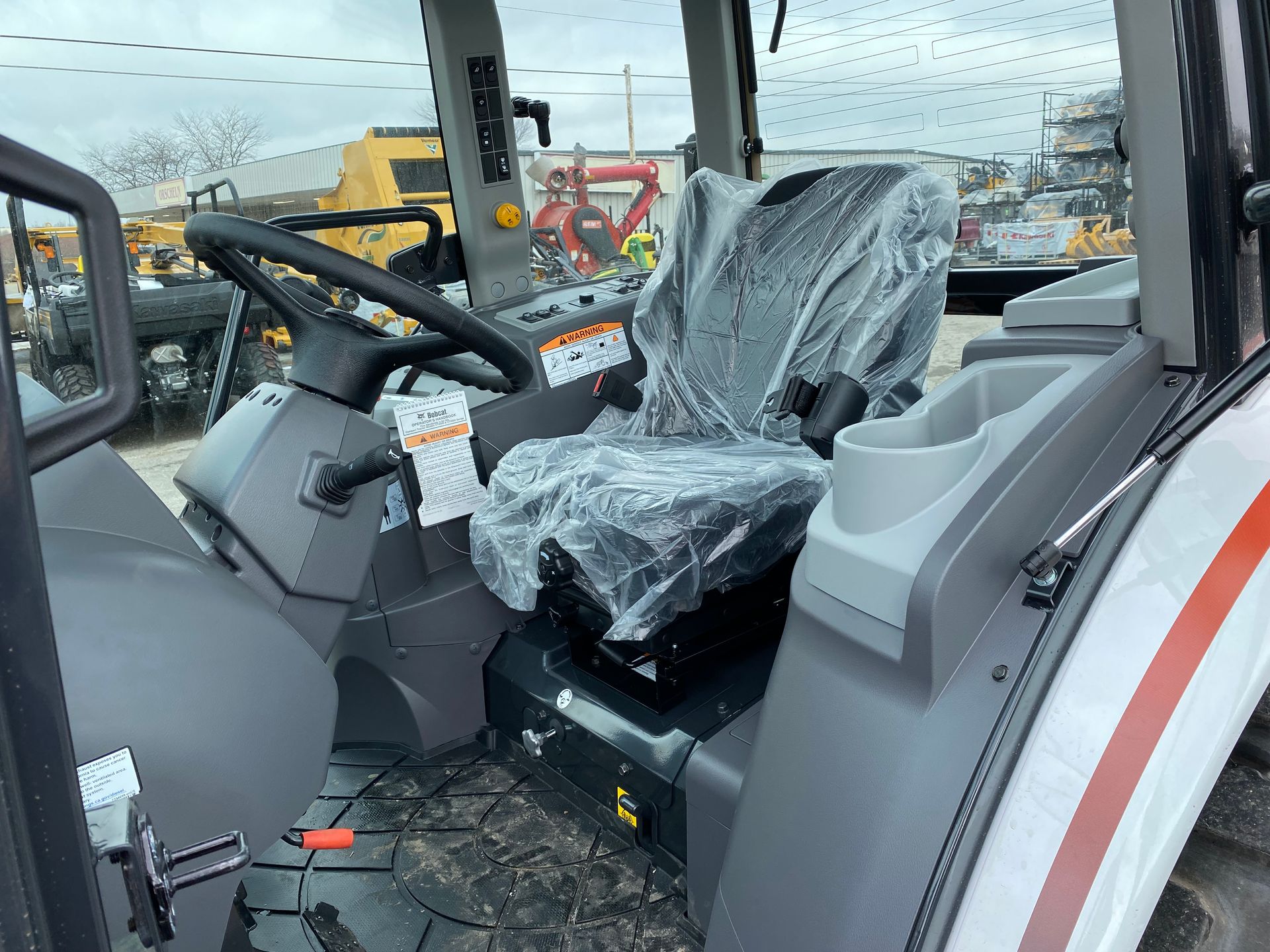 Interior view of tractor cab, black steering wheel, gray seat, protective plastic wrap, outside setting.