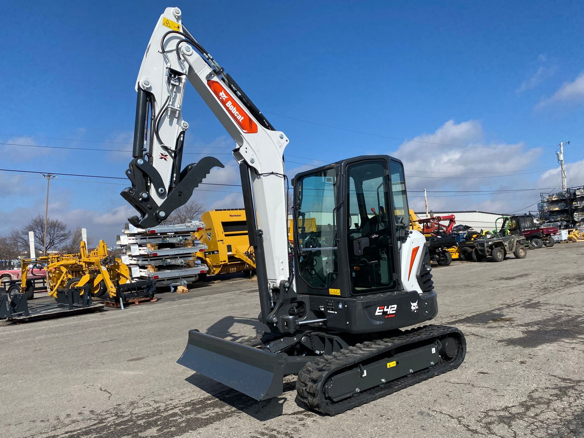 Bobcat excavator with raised arm and grapple attachment, outdoors on pavement. White and black with orange logo.