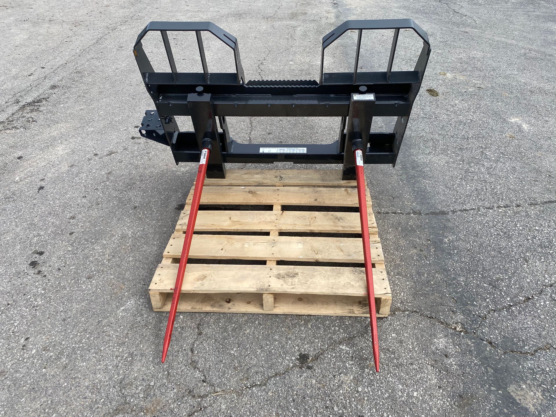 Black and red bale spear attachment resting on a wooden pallet outdoors on a gray surface.