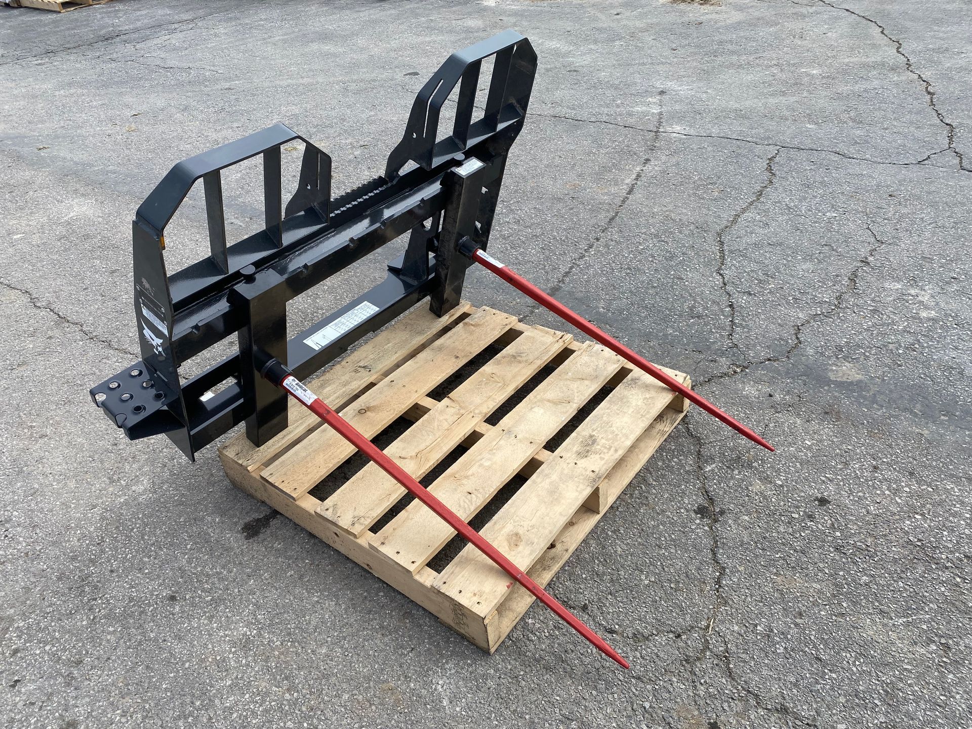 A black hay bale spear attachment resting on a wooden pallet; two red spears extend forward.