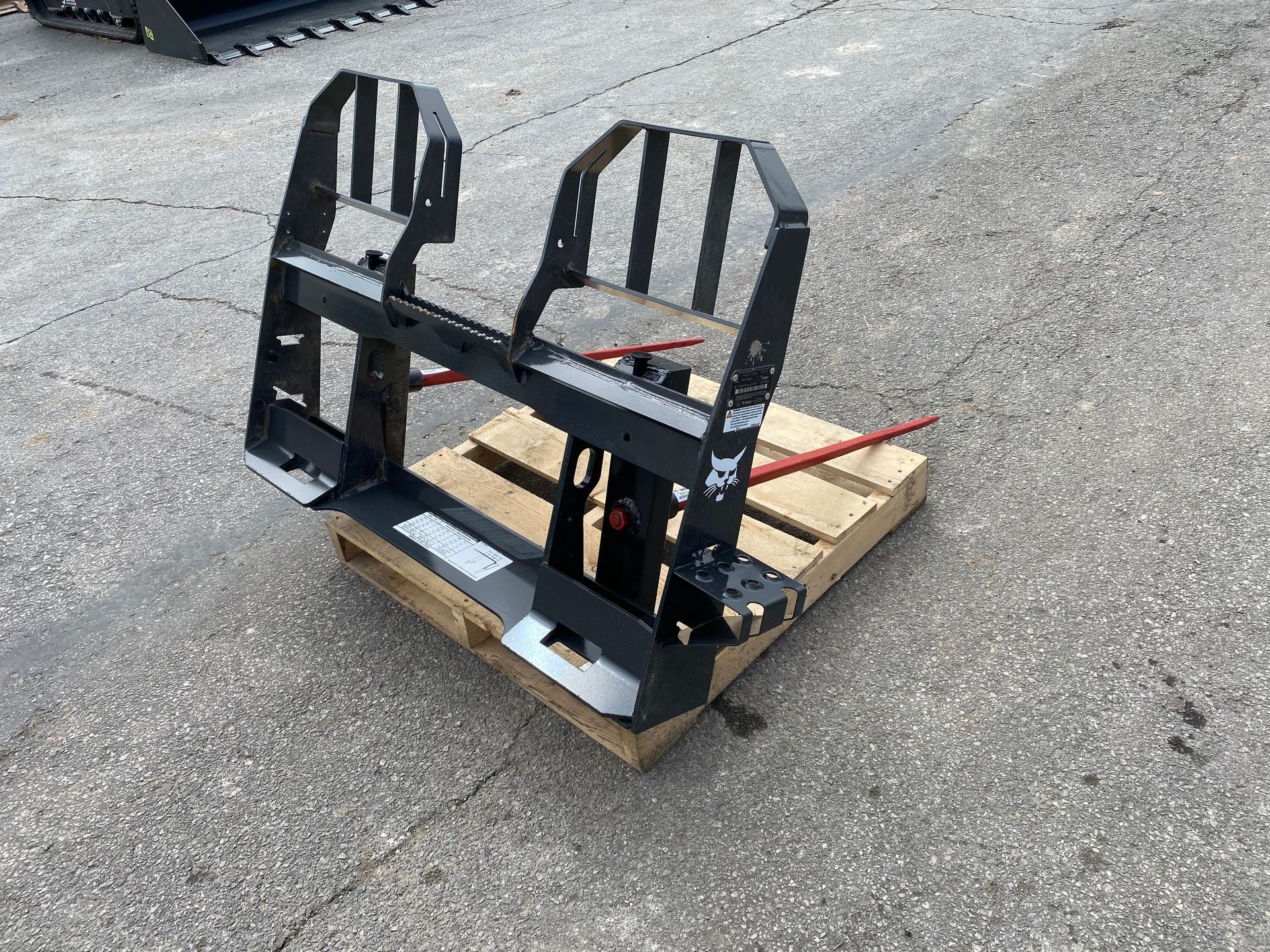 Black Bobcat pallet fork attachment on a wooden pallet, outdoors on pavement.
