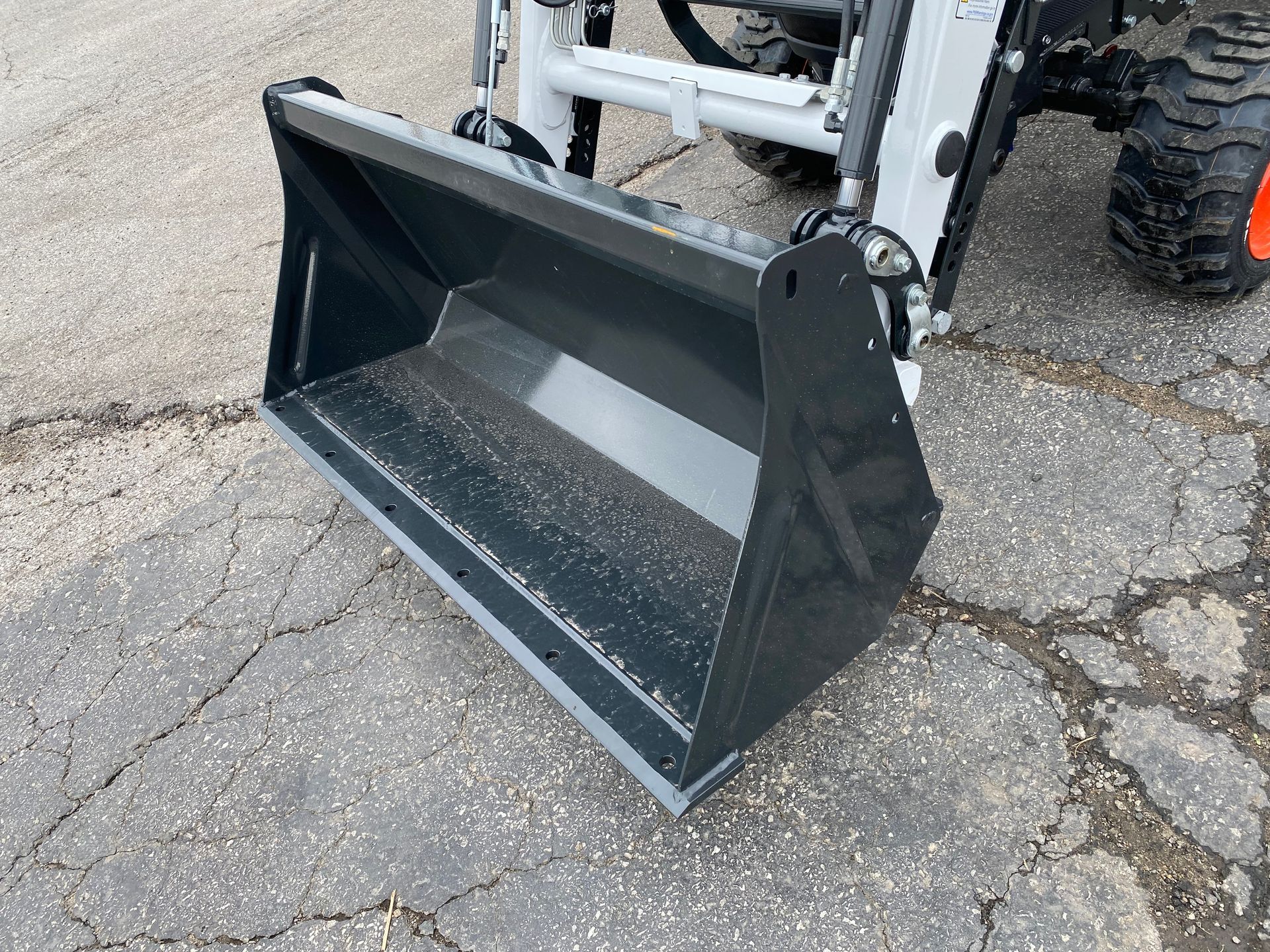 Black tractor bucket attached to a white tractor, resting on gray pavement.