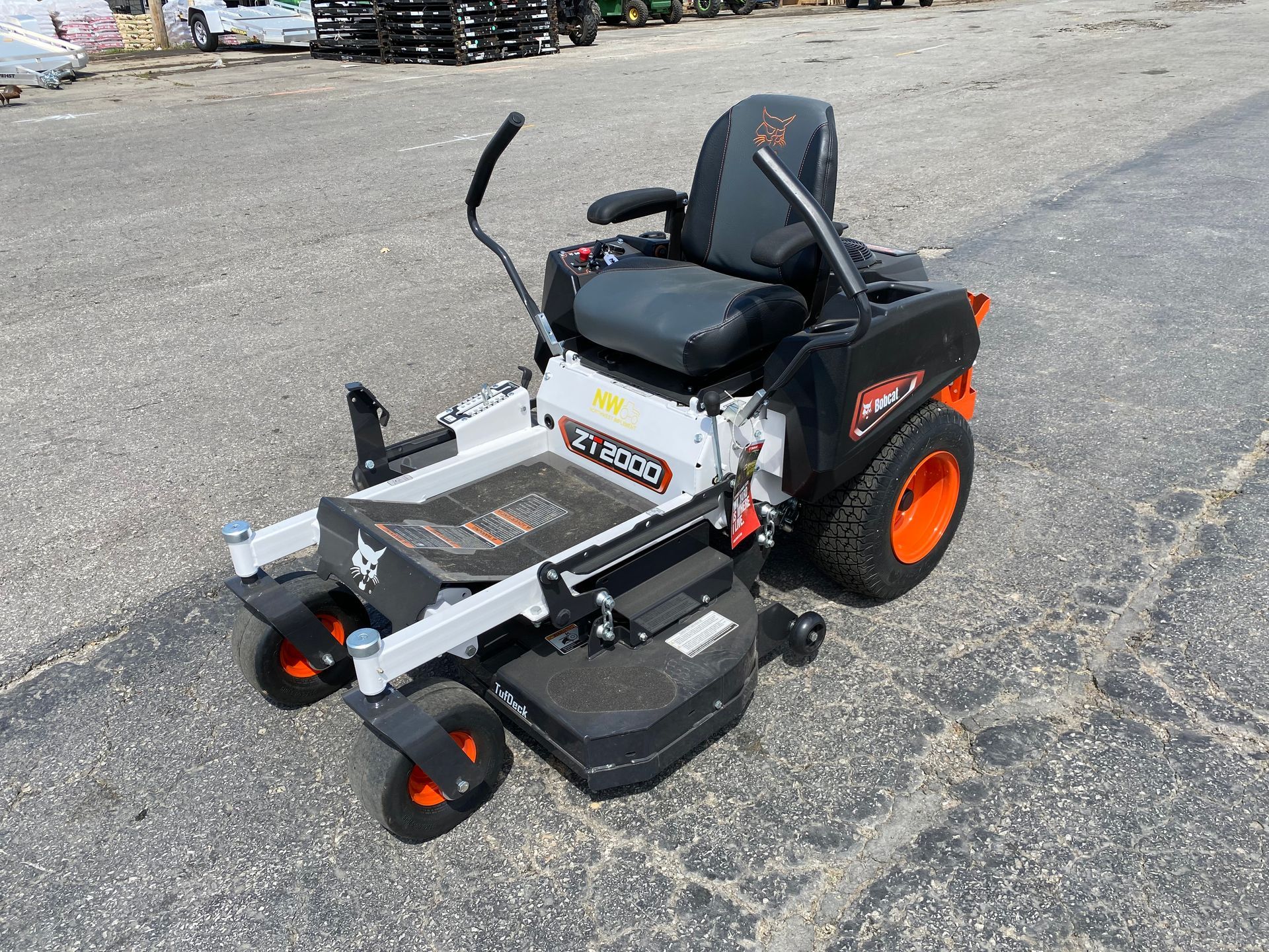 Bobcat zero-turn lawn mower on asphalt, black frame, orange wheels, gray deck and seat, in a sunny outdoor setting.