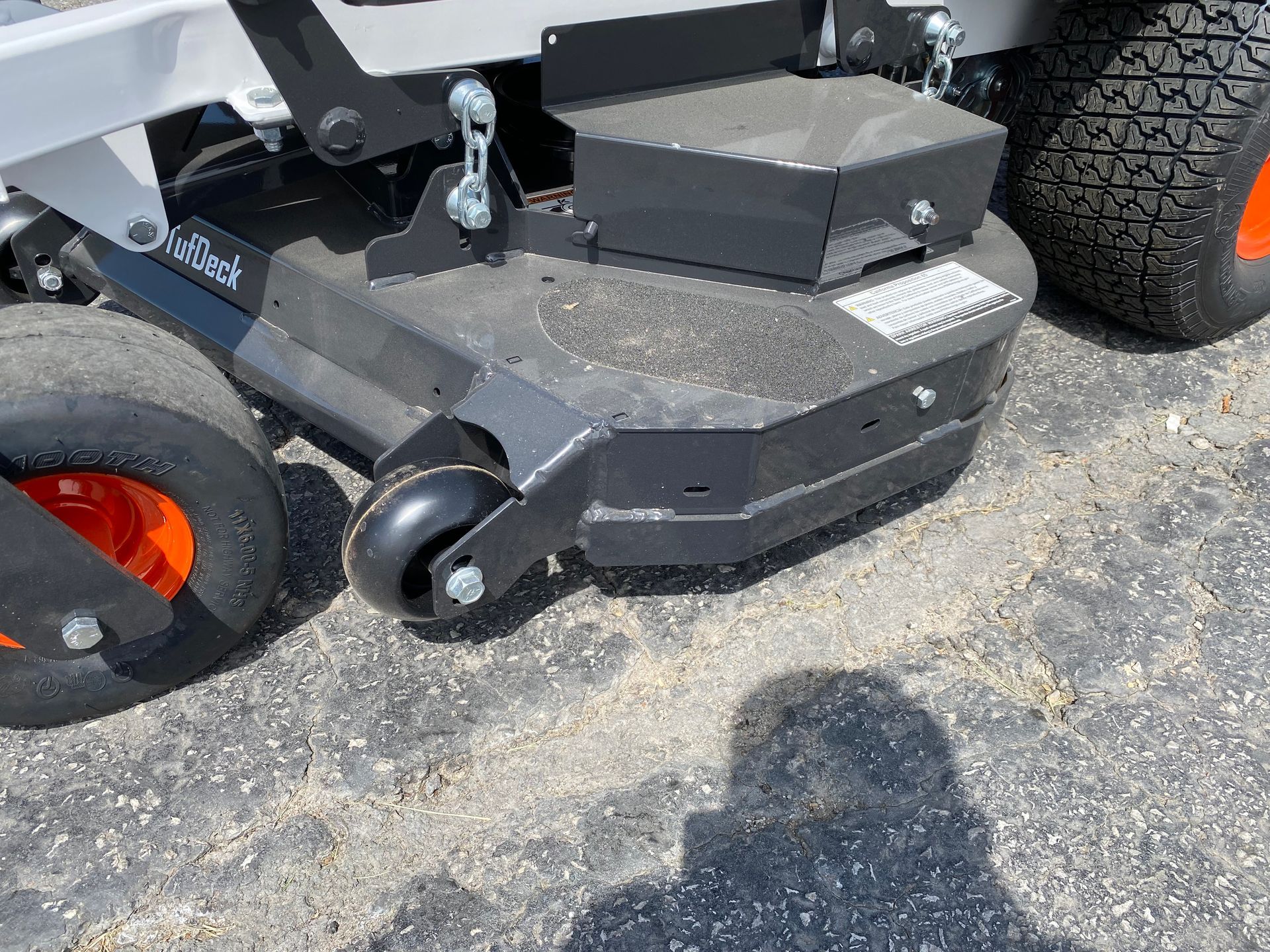 Black and gray zero-turn mower deck with orange wheels on asphalt.