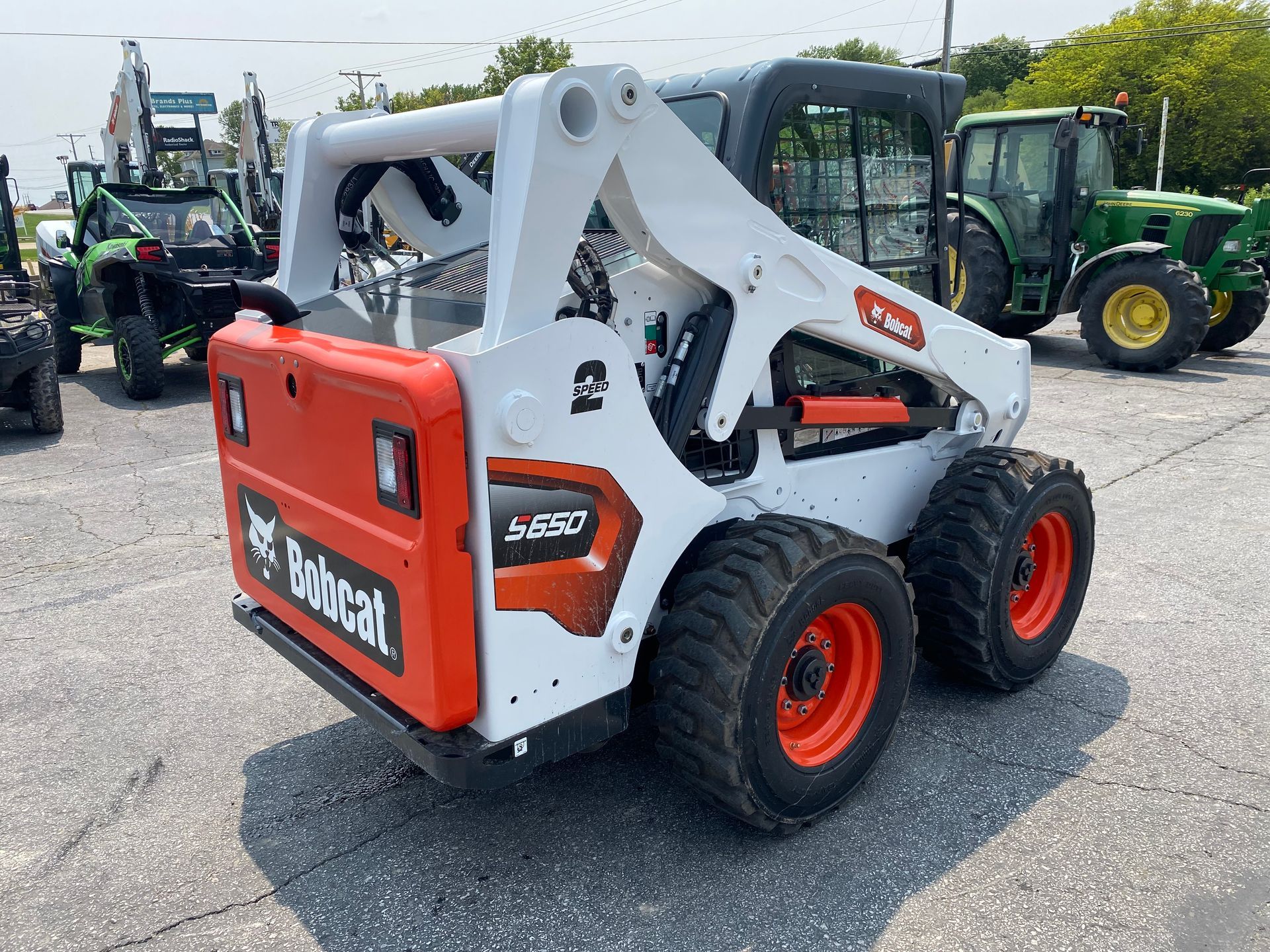 White and orange Bobcat S650 skid-steer loader parked on pavement, with a green tractor in the background.