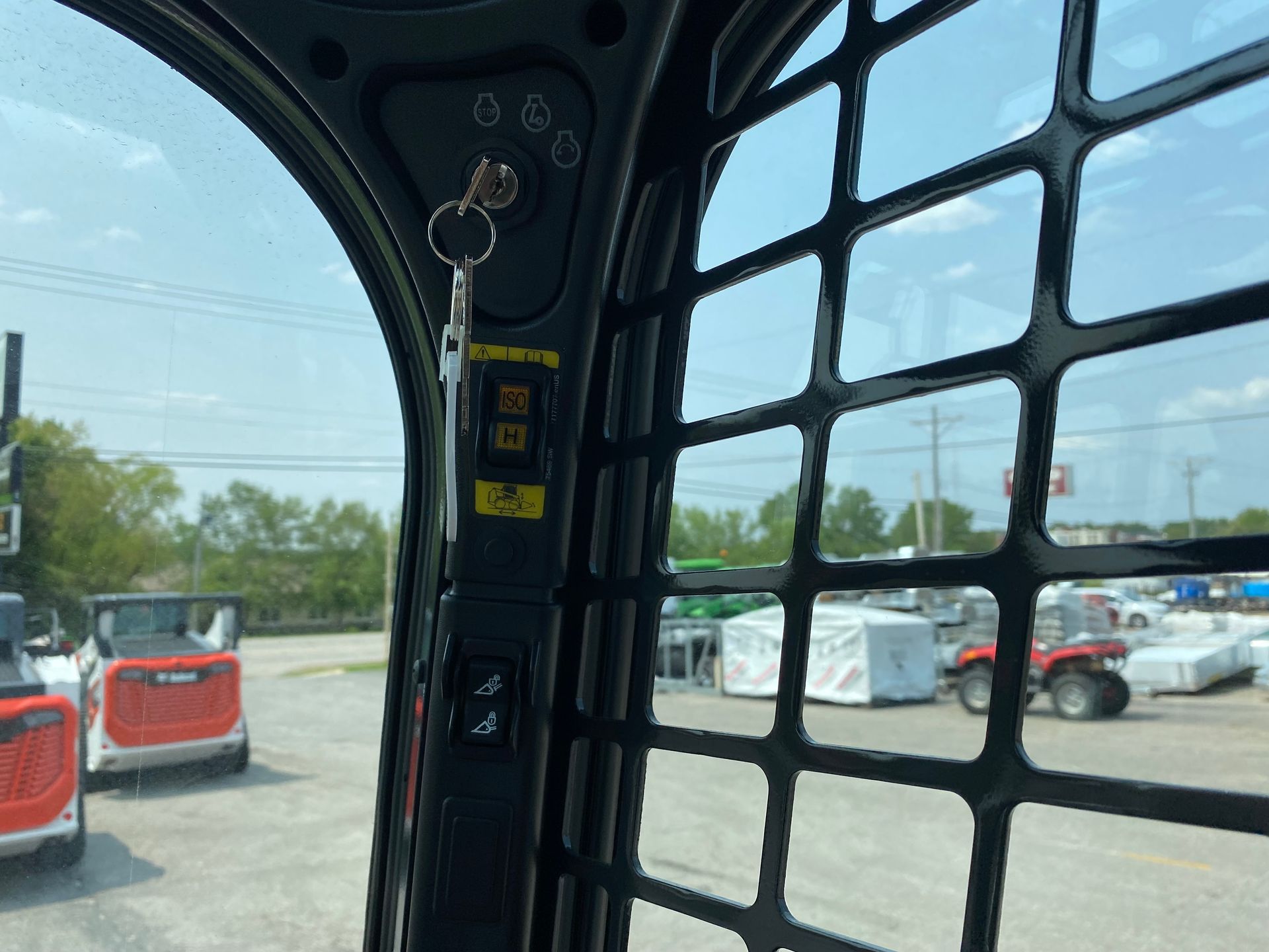 Interior of a Bobcat skid steer cab, showing keys, controls, and a view of other Bobcats and a vehicle outside.