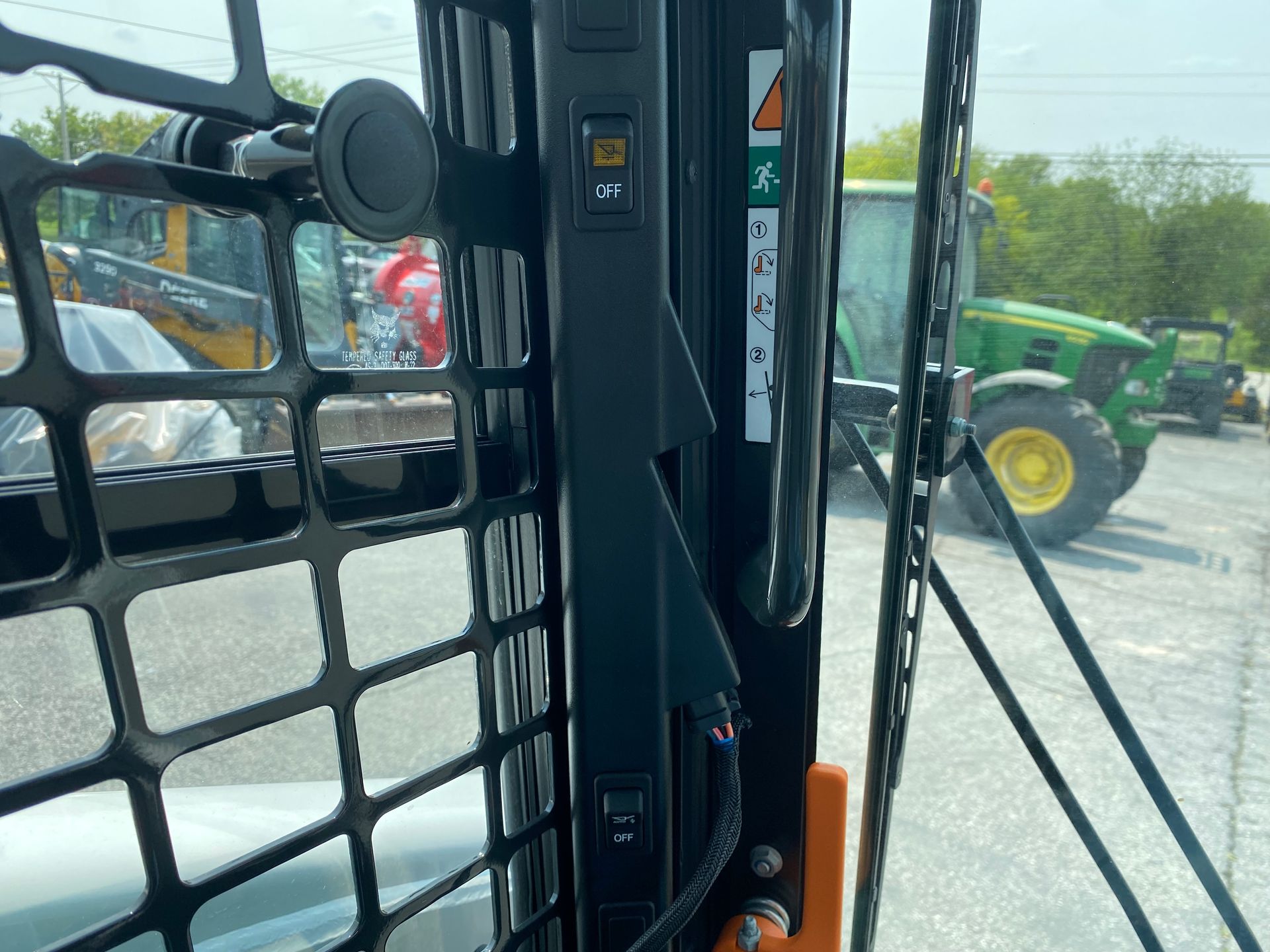 Interior of a skid steer cab with controls, safety symbols, and a partially visible John Deere tractor in the background.