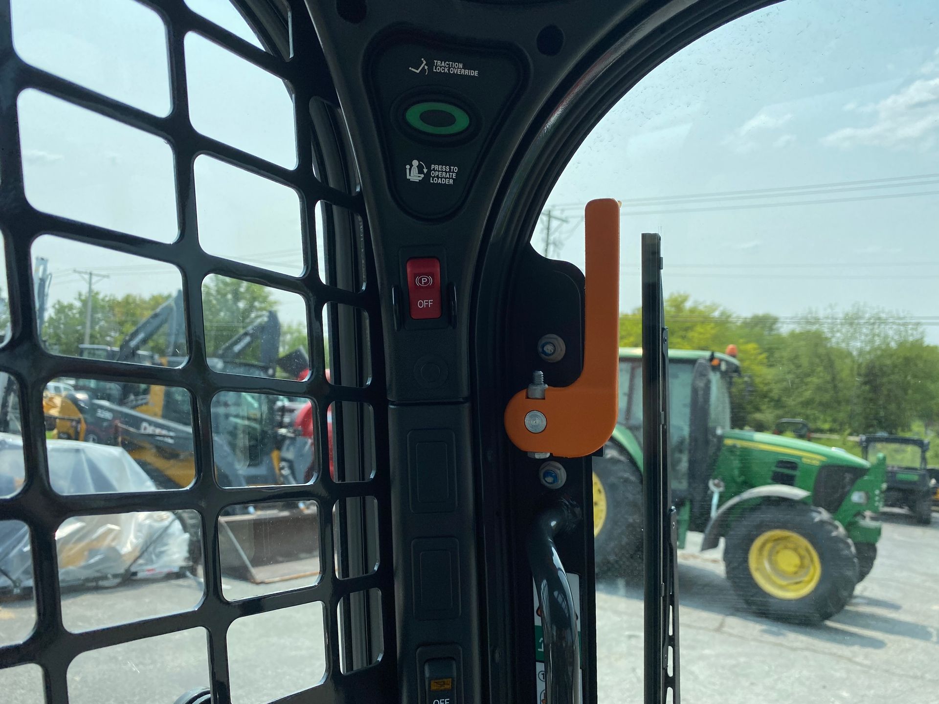 Interior view of a skid steer cab, showing control panel, safety labels, and a partial view of a green tractor outside.