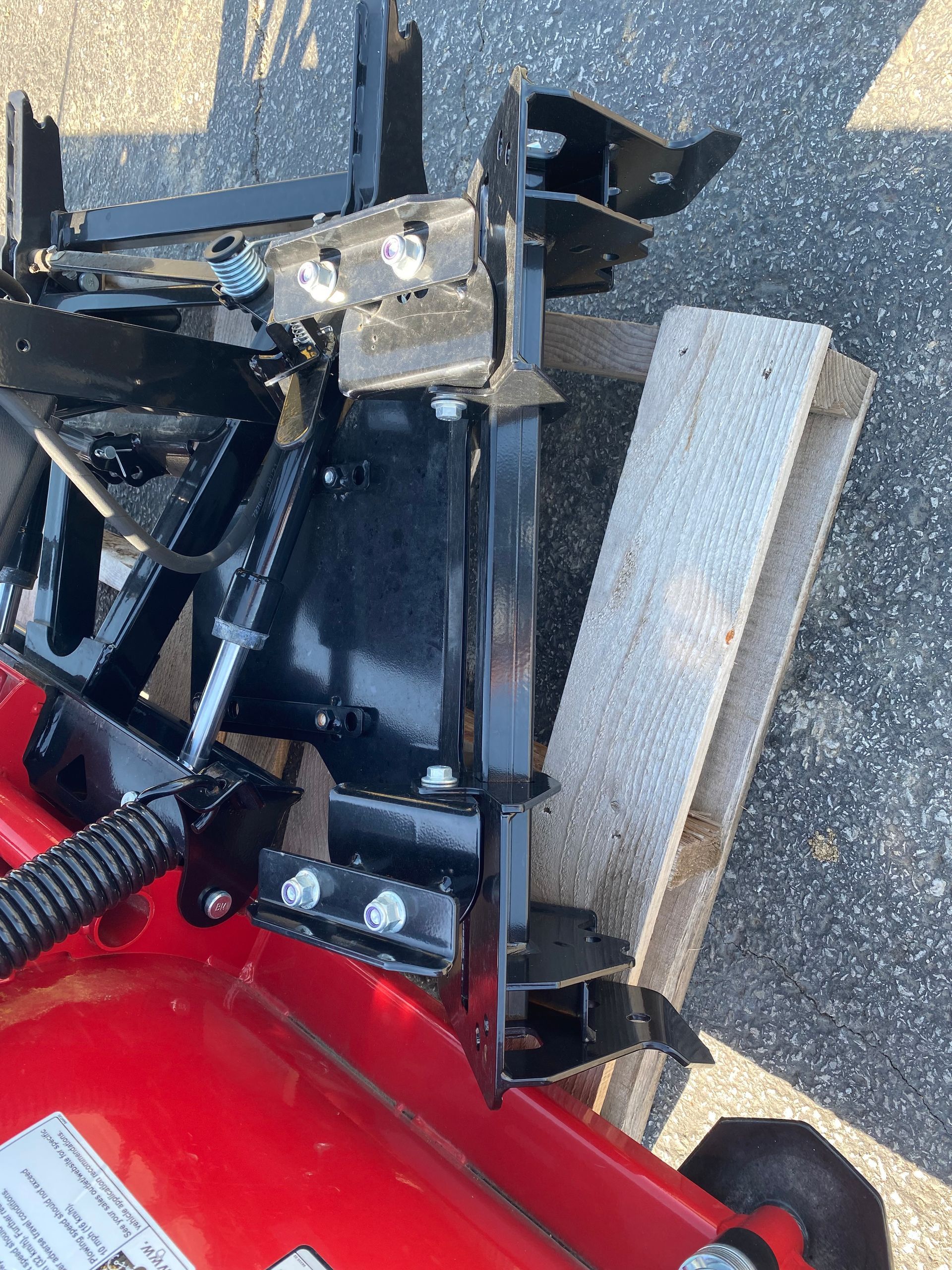 Red blade with a black mounting attachment, propped up by a wooden plank on asphalt.