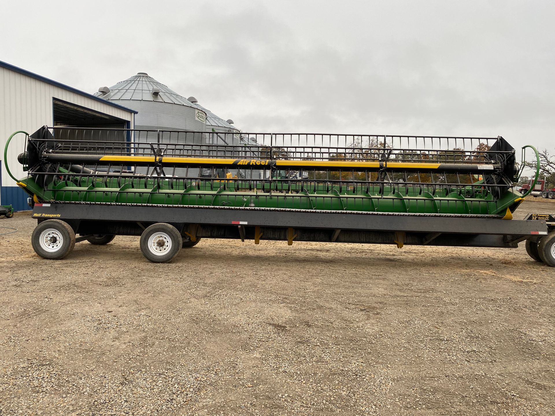 Green and black combine header on a black trailer, parked outside a building and grain silo.