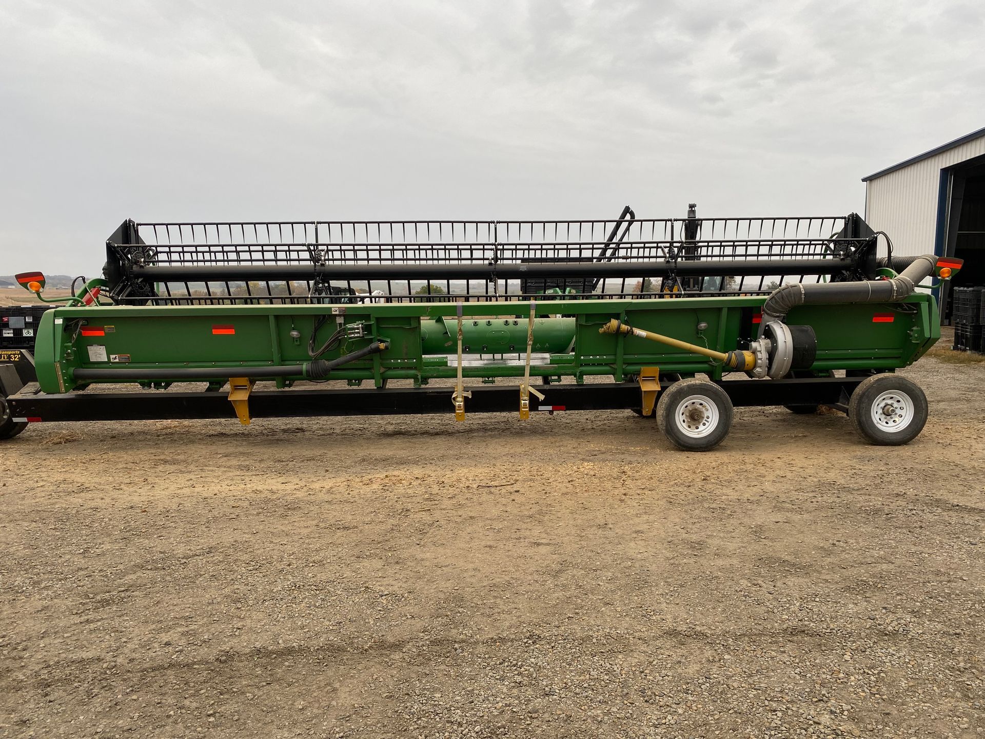 Green and black agricultural header on a trailer, parked on gravel in front of a building.