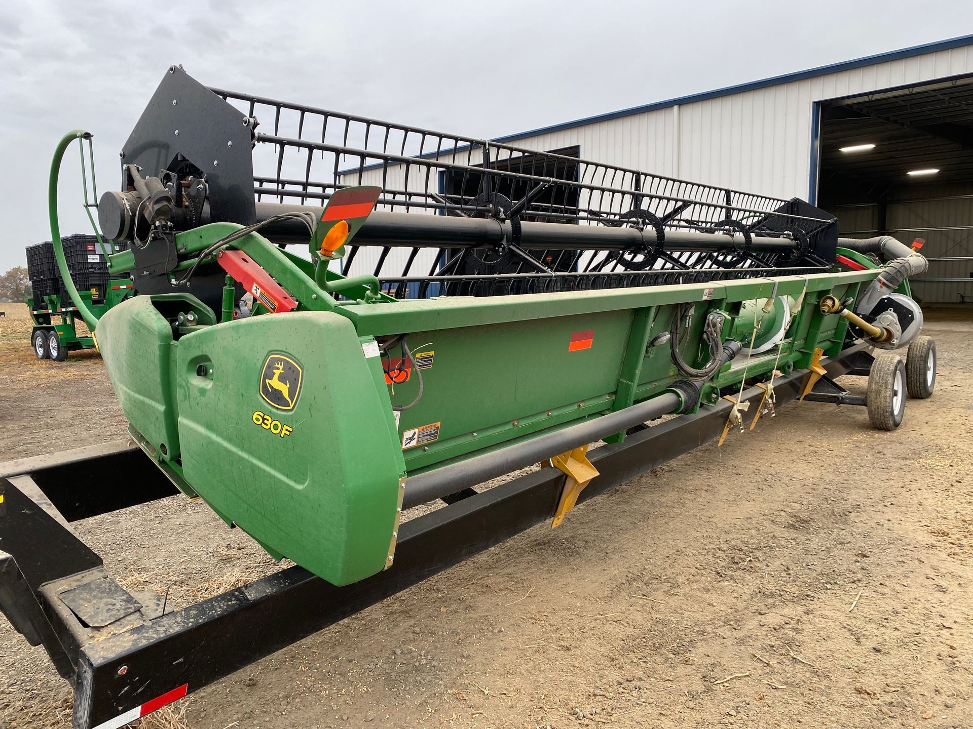 John Deere combine header on a black trailer, parked outside a building on a cloudy day.