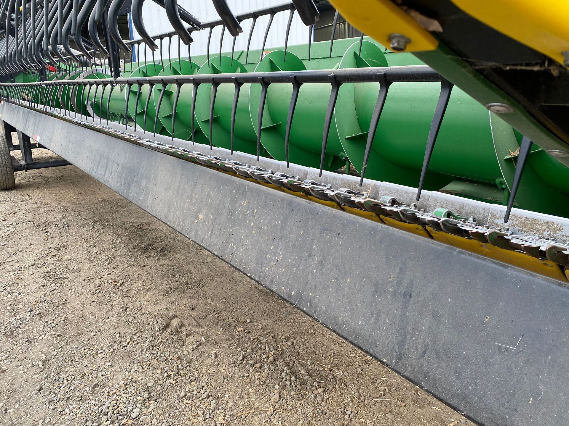 Close-up of a combine header. Dark bar with tines, green cylinders in background.