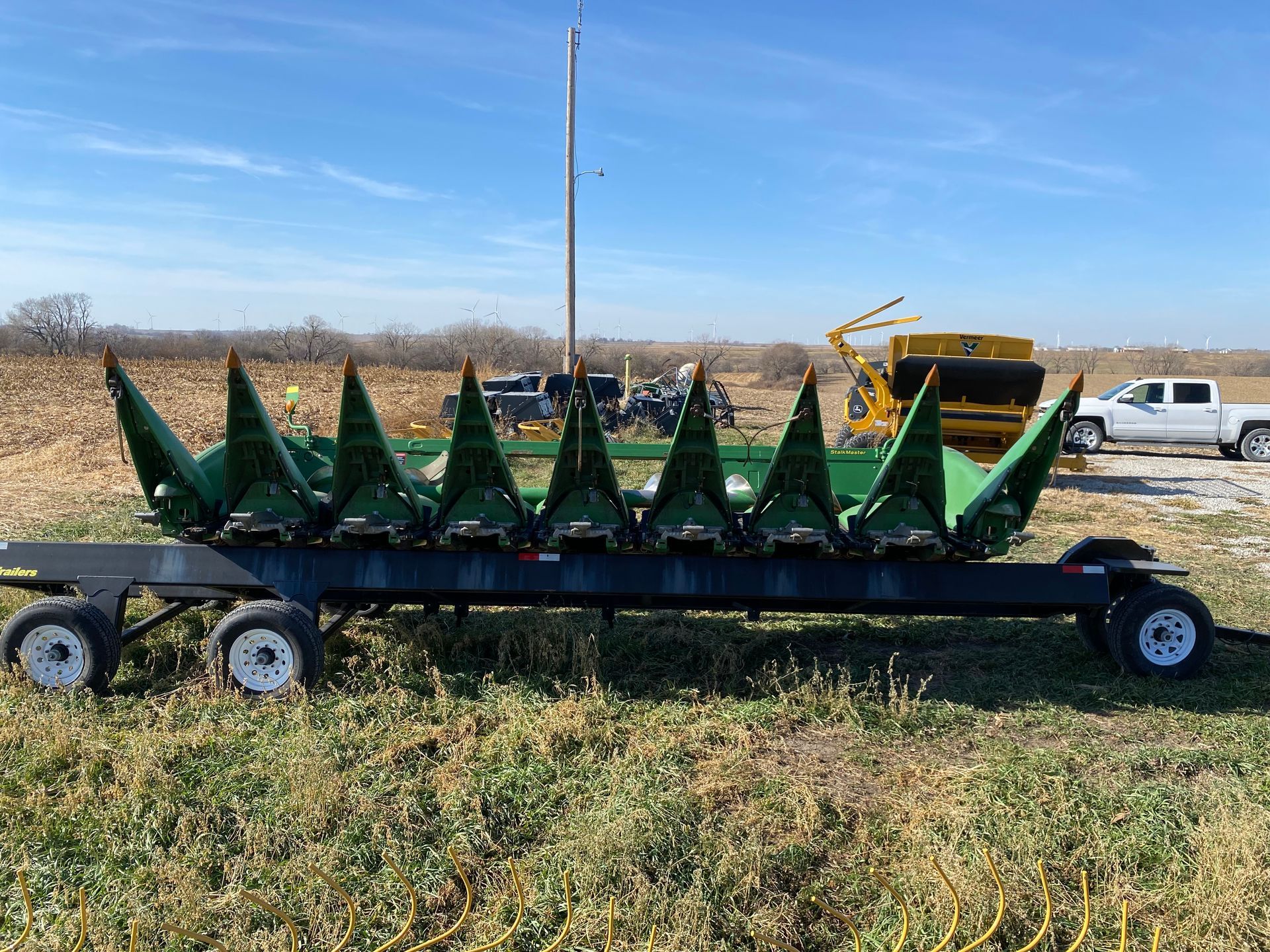 A corn head on a trailer, outdoors, with a truck and farm equipment in the background.
