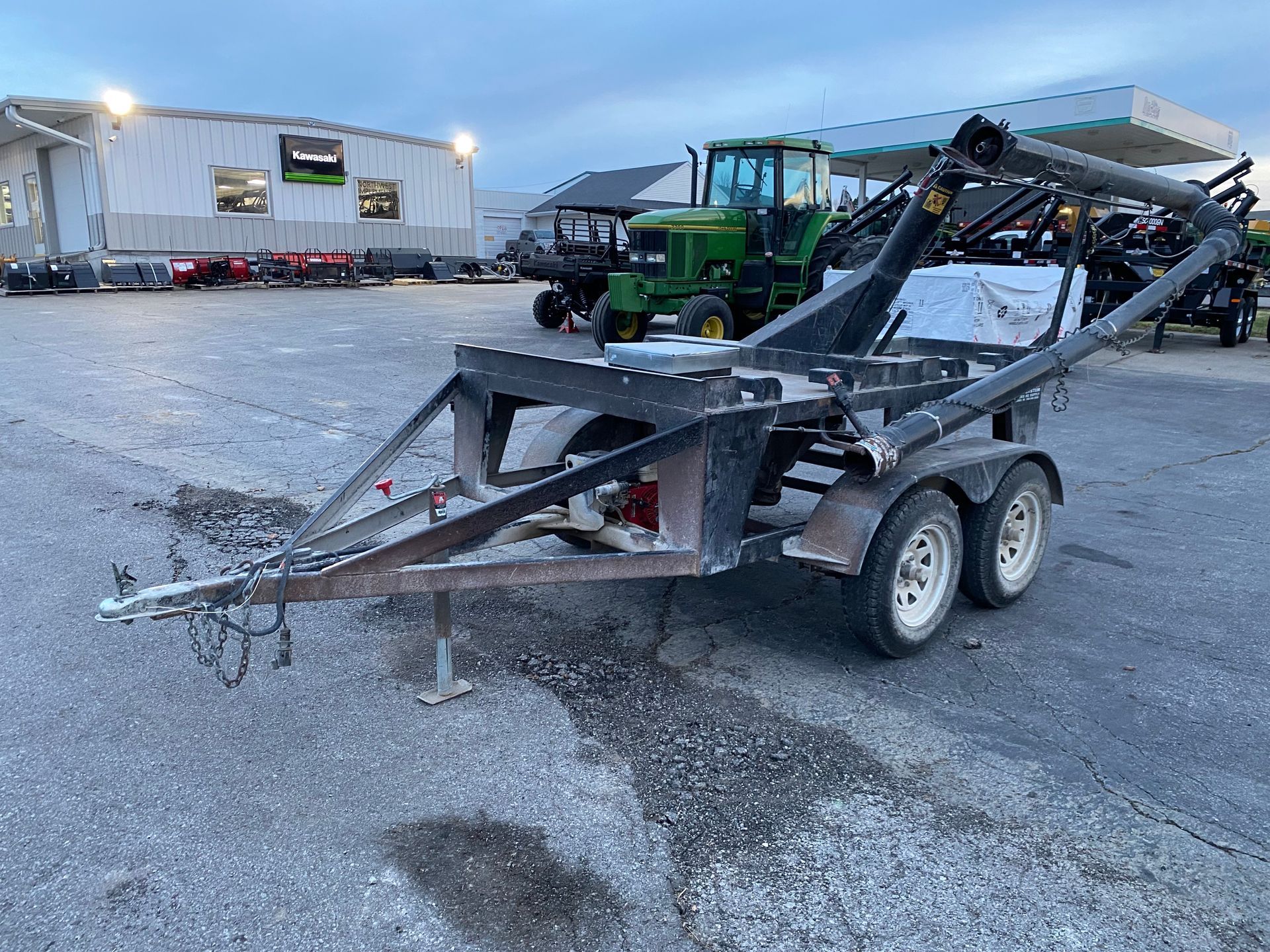 Black trailer with dual wheels, a frame, and an arm, parked on asphalt with a tractor in the background.