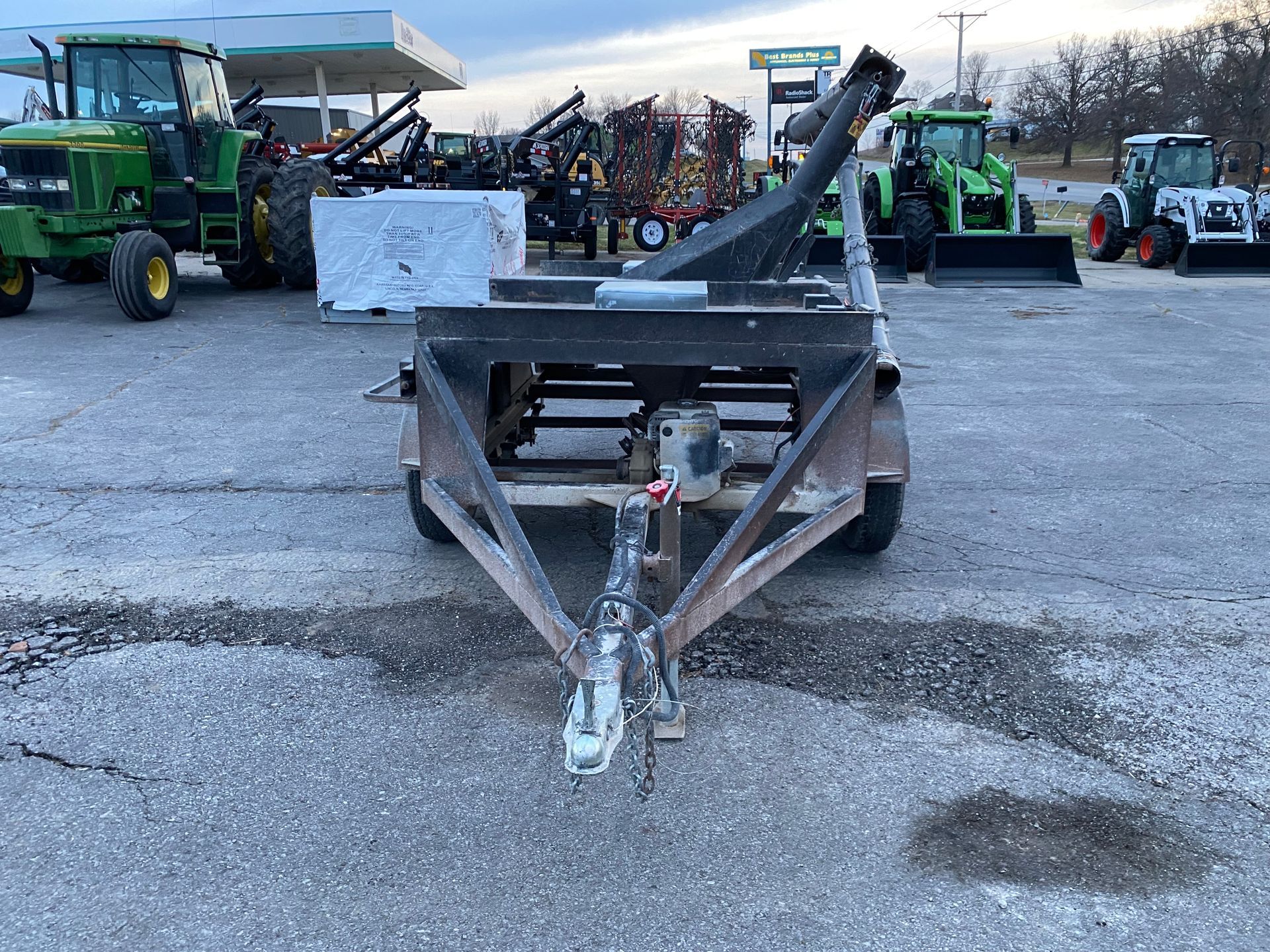A utility trailer with a long angled arm, parked in a lot with tractors and a gas station.