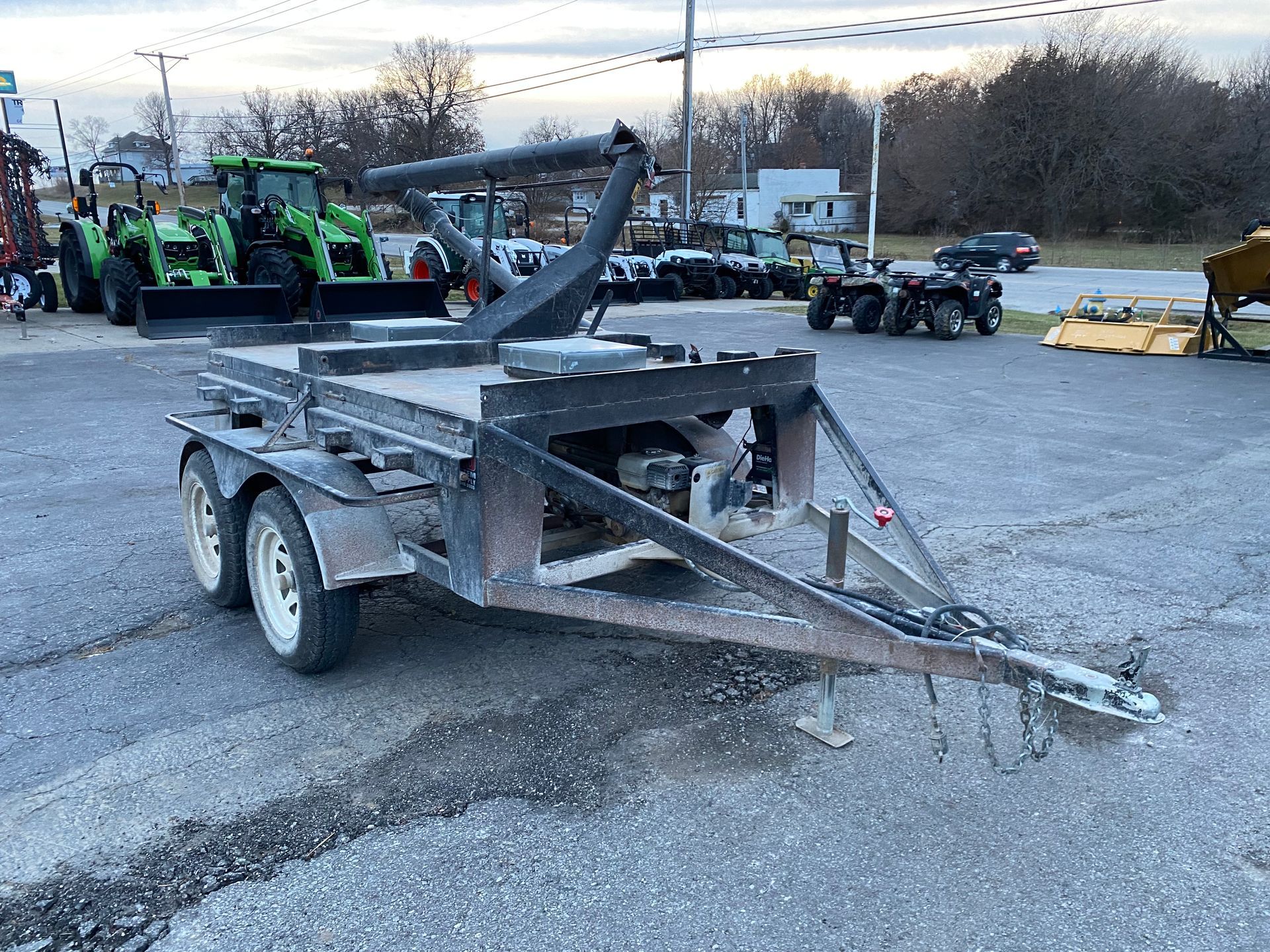 Utility trailer with a built-in log splitter, parked on asphalt. Other equipment and vehicles are visible in the background.