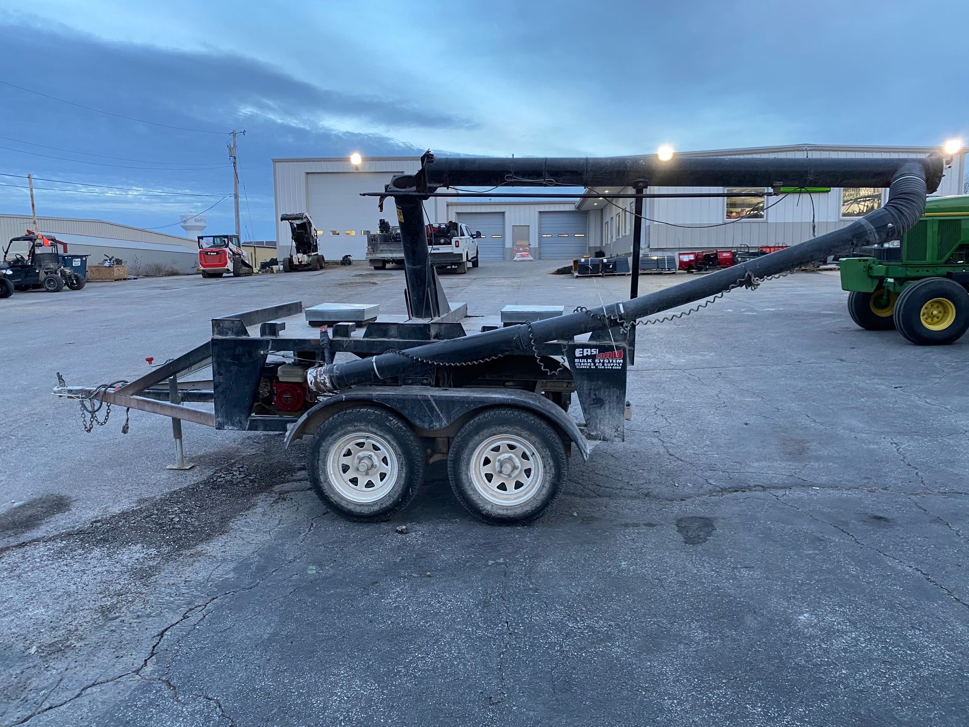 Black trailer with auger, parked on asphalt; industrial setting, evening.