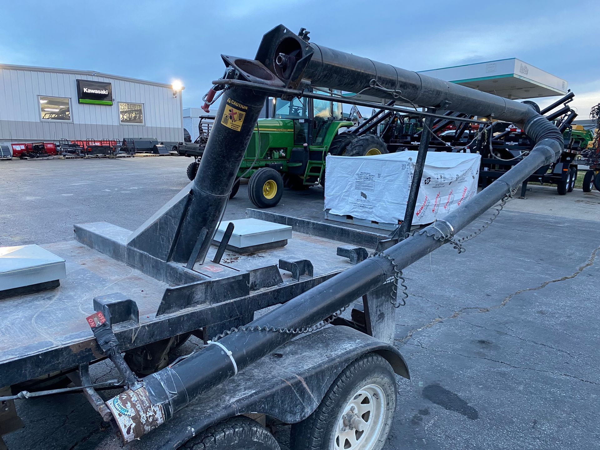 Black grain auger on a trailer, with tractors and a gas station in the background.