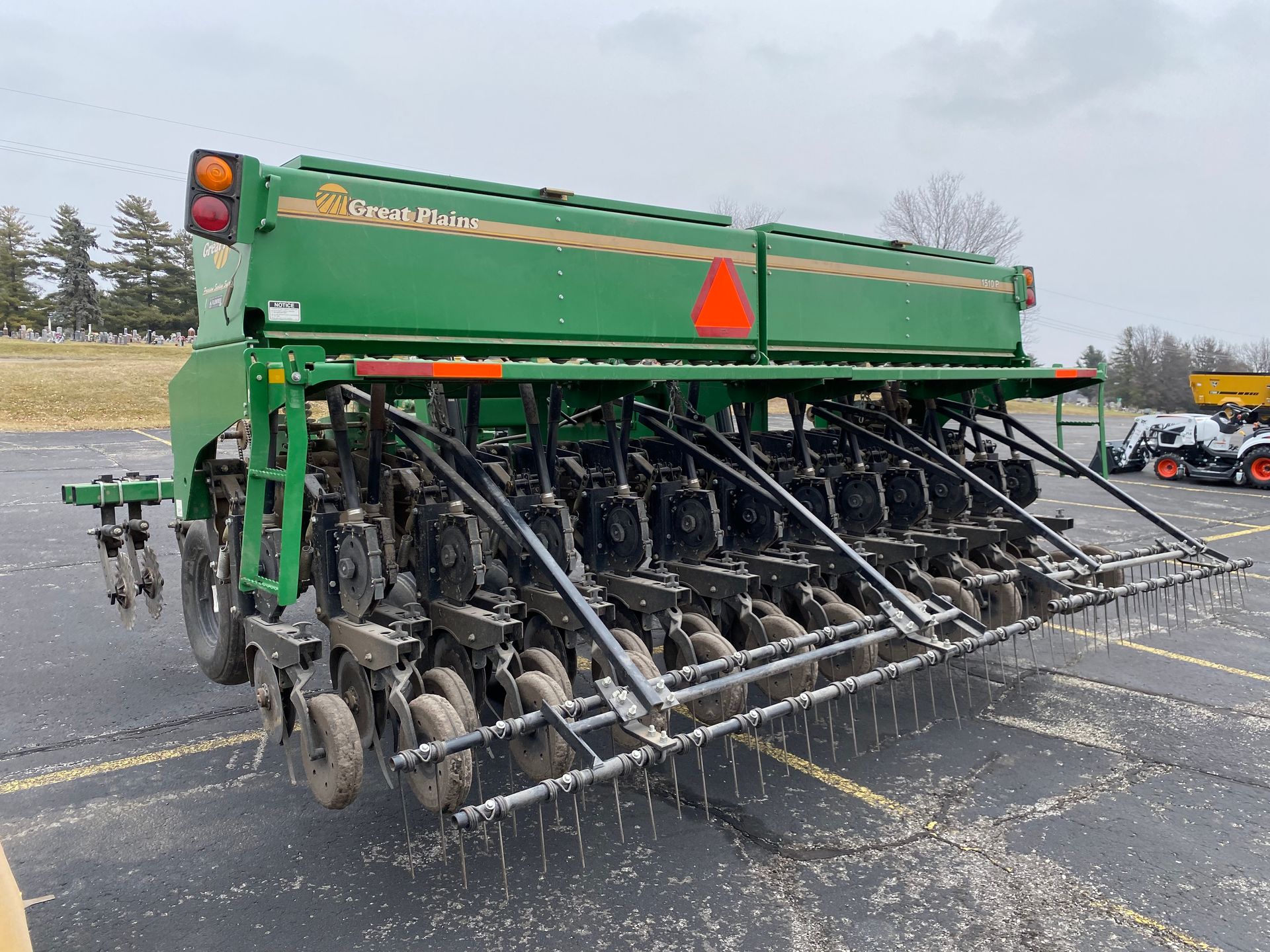 Green agricultural grain drill parked on asphalt, cloudy day, back angled shot.