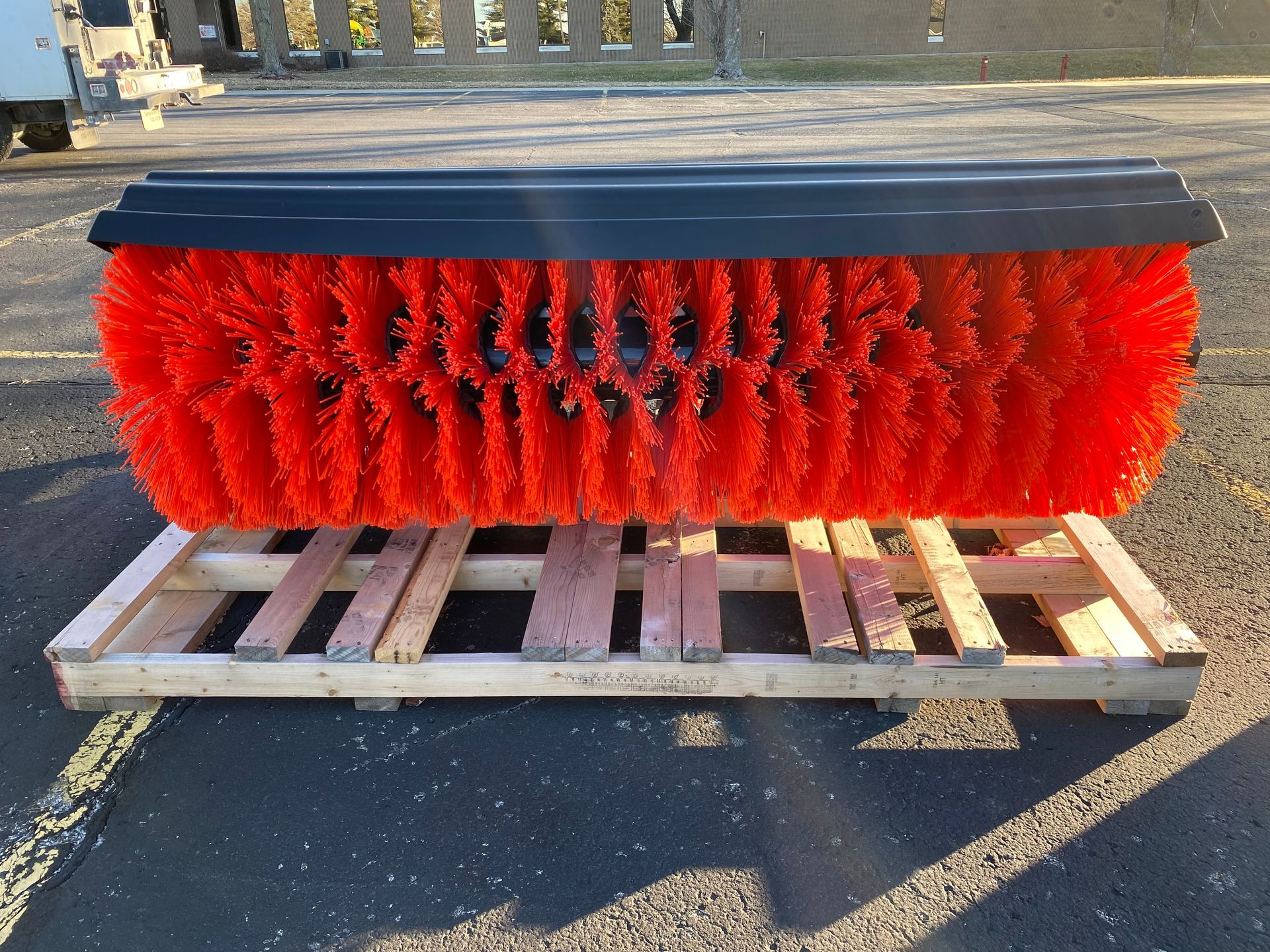 Red and black industrial street sweeper brush on a wooden pallet outdoors.