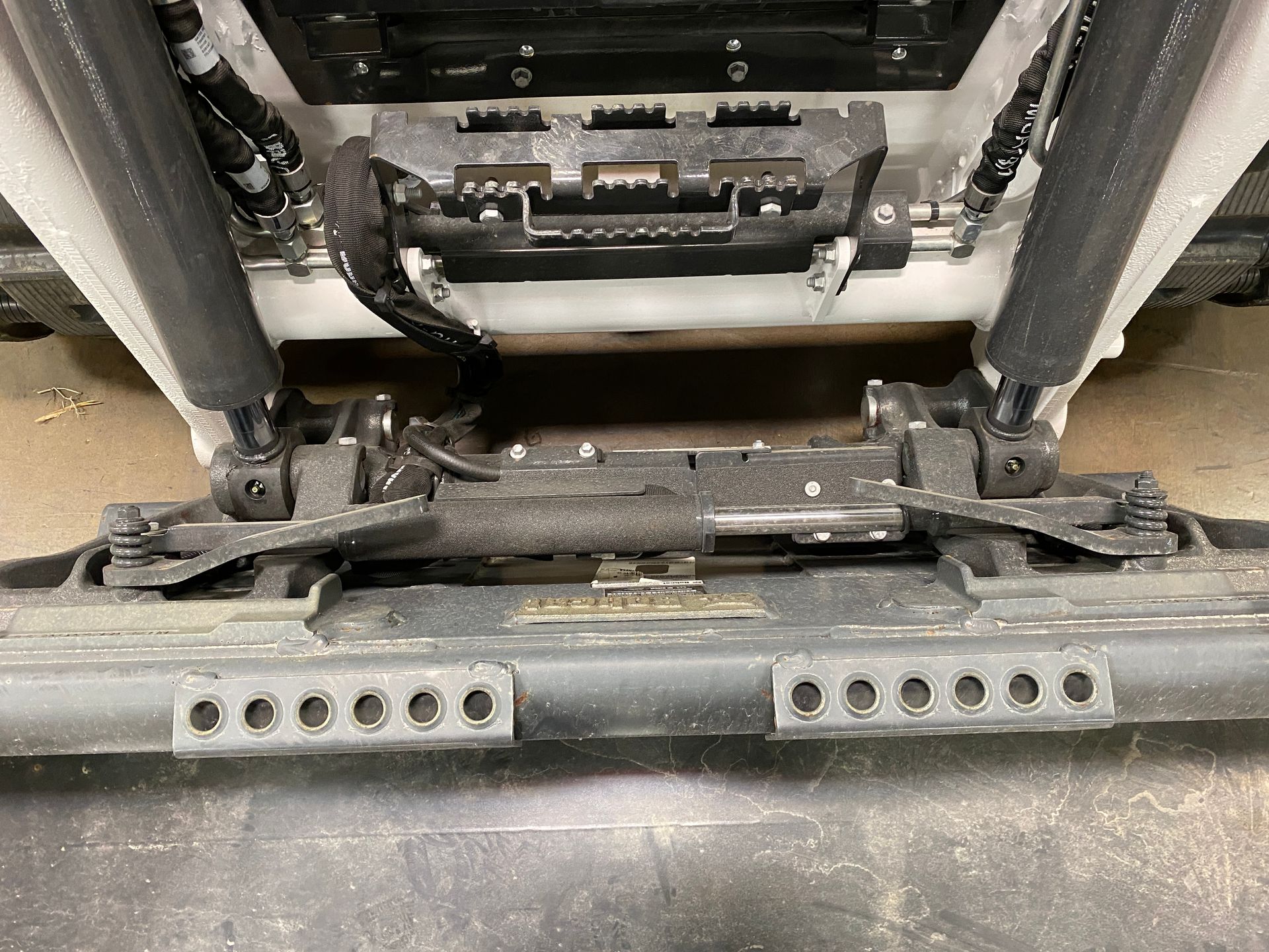 Underside view of a black snowplow with two cylinders, metal frame, and multiple bolt holes.