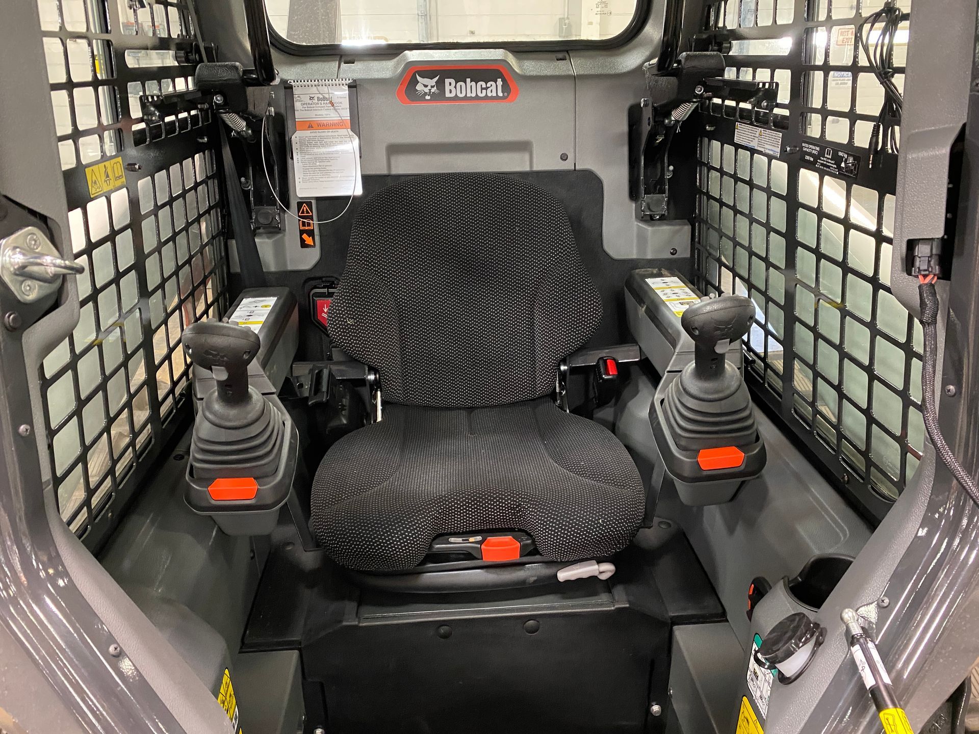 Interior of a Bobcat skid-steer loader showing the operator's seat, joysticks, and safety cage.