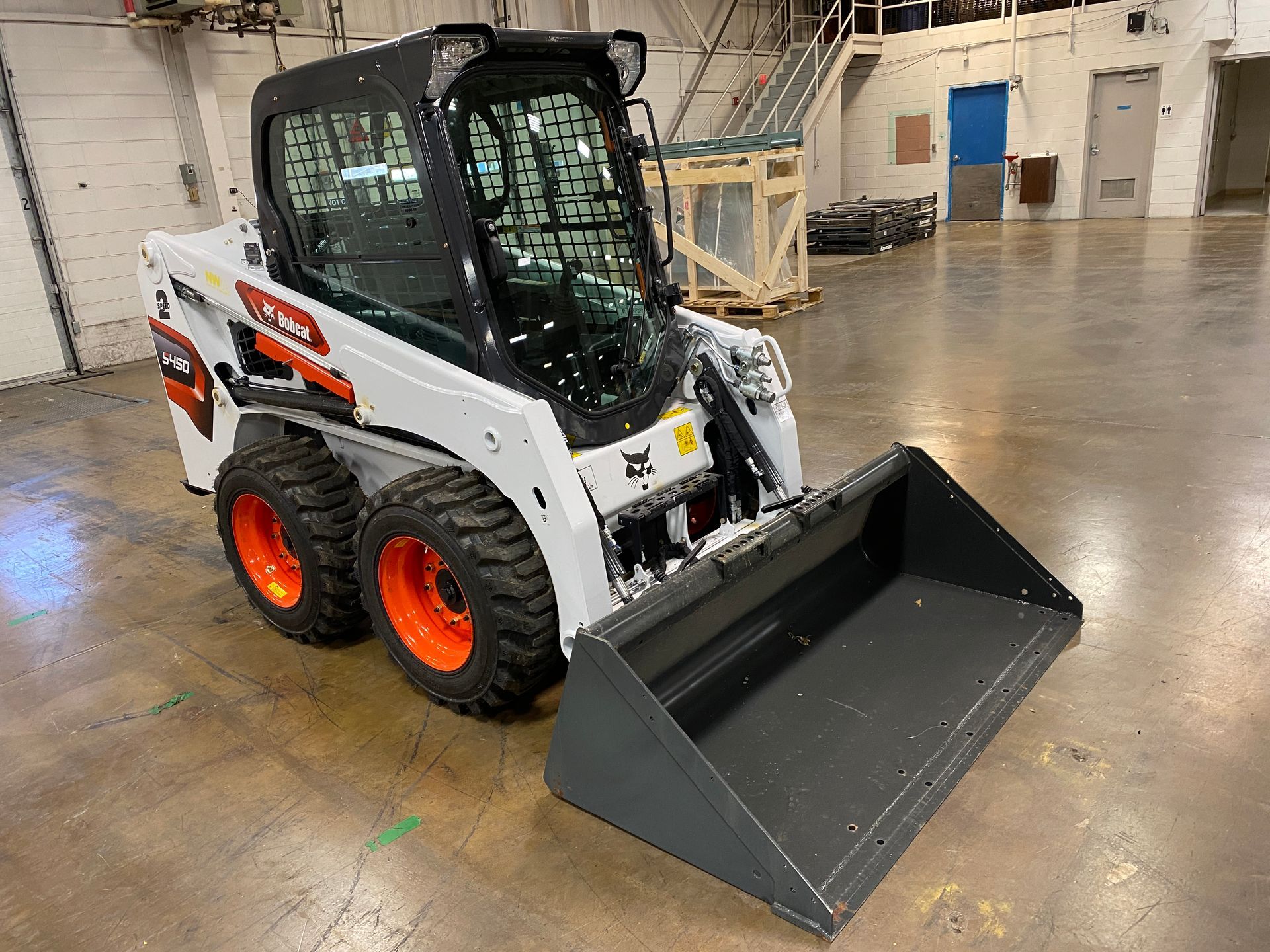 White and orange Bobcat skid-steer loader with a bucket indoors, on a concrete floor.
