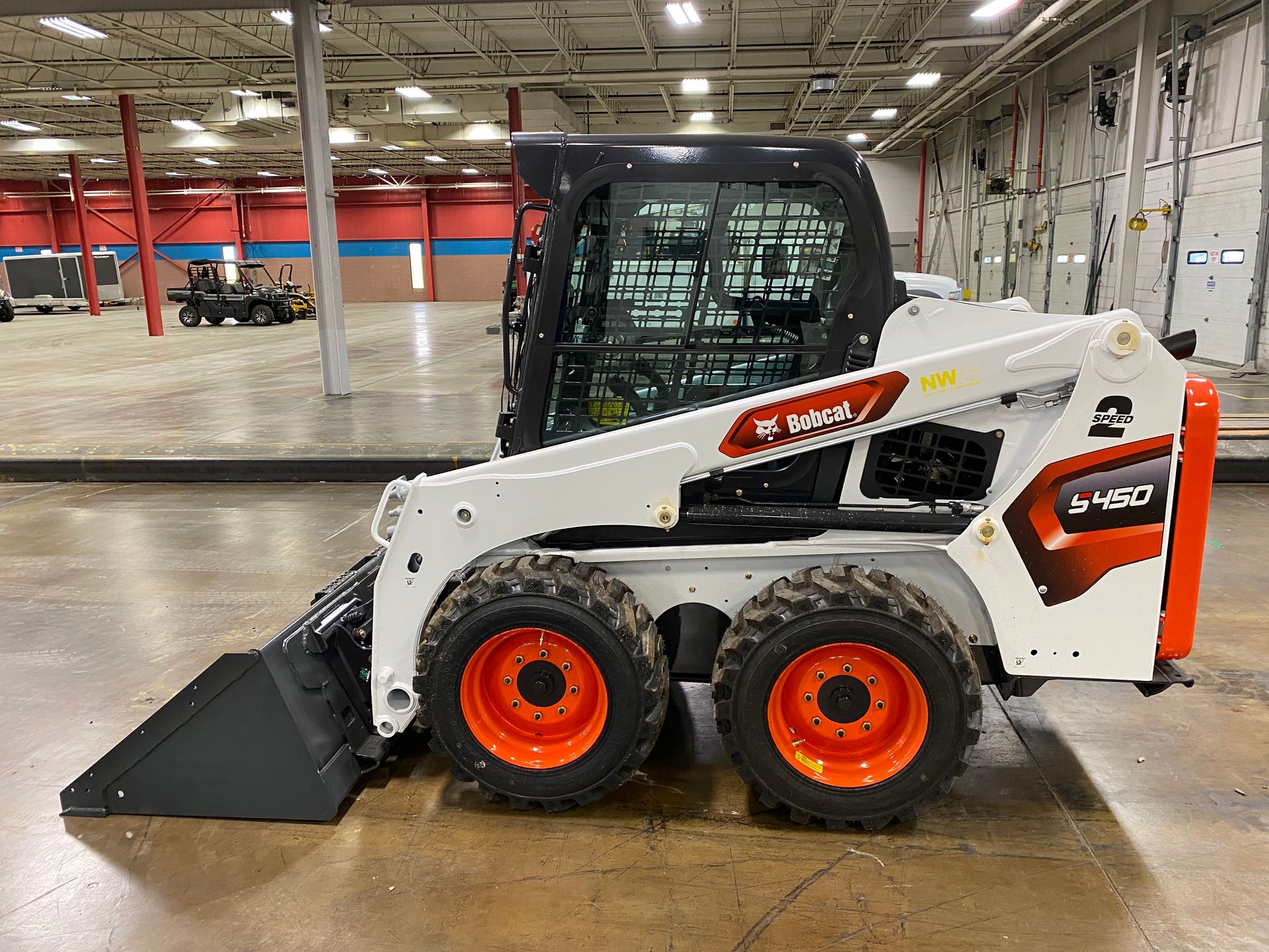 Bobcat S450 skid-steer loader, white with orange wheels, bucket attached, indoors.