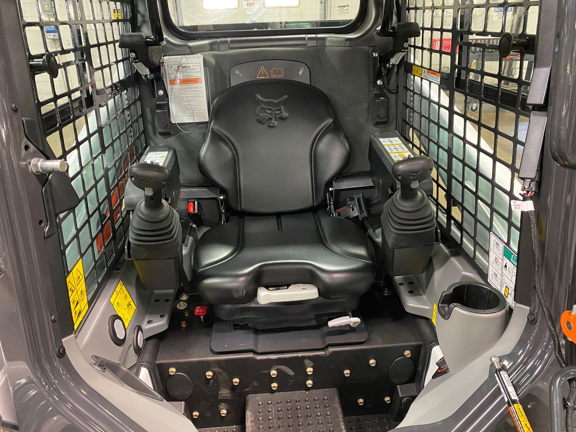 Interior of a Bobcat skid steer: black seat, joystick controls, protective side grilles, cup holder.