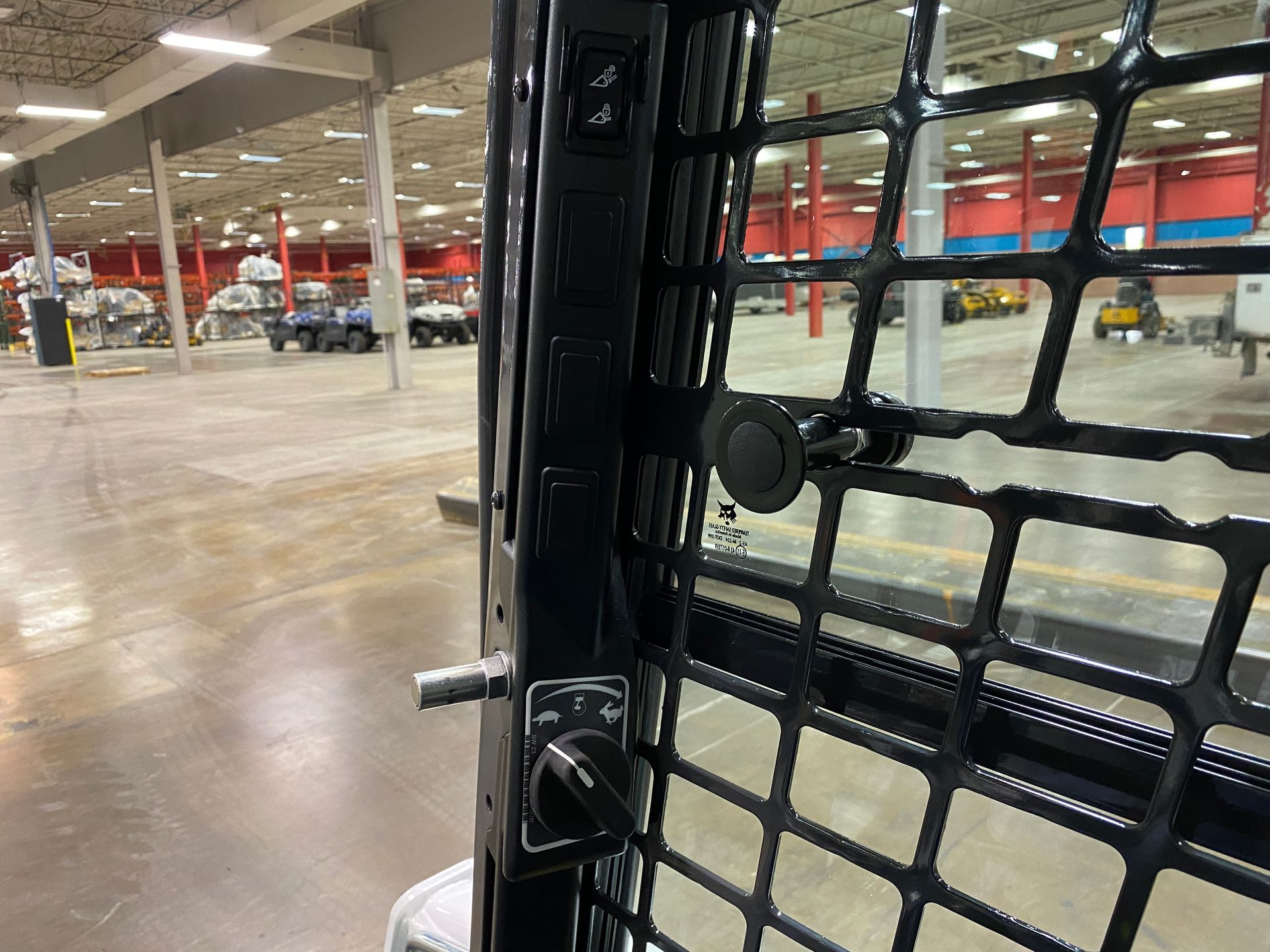 Interior view of a skid steer cab, showing the safety grill, door latch, and warehouse setting.