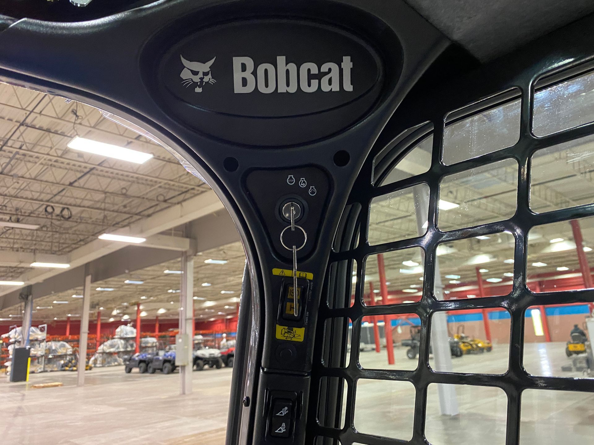 Bobcat skid-steer interior view, key in ignition. Overhead logo, safety cage, and warehouse background visible.