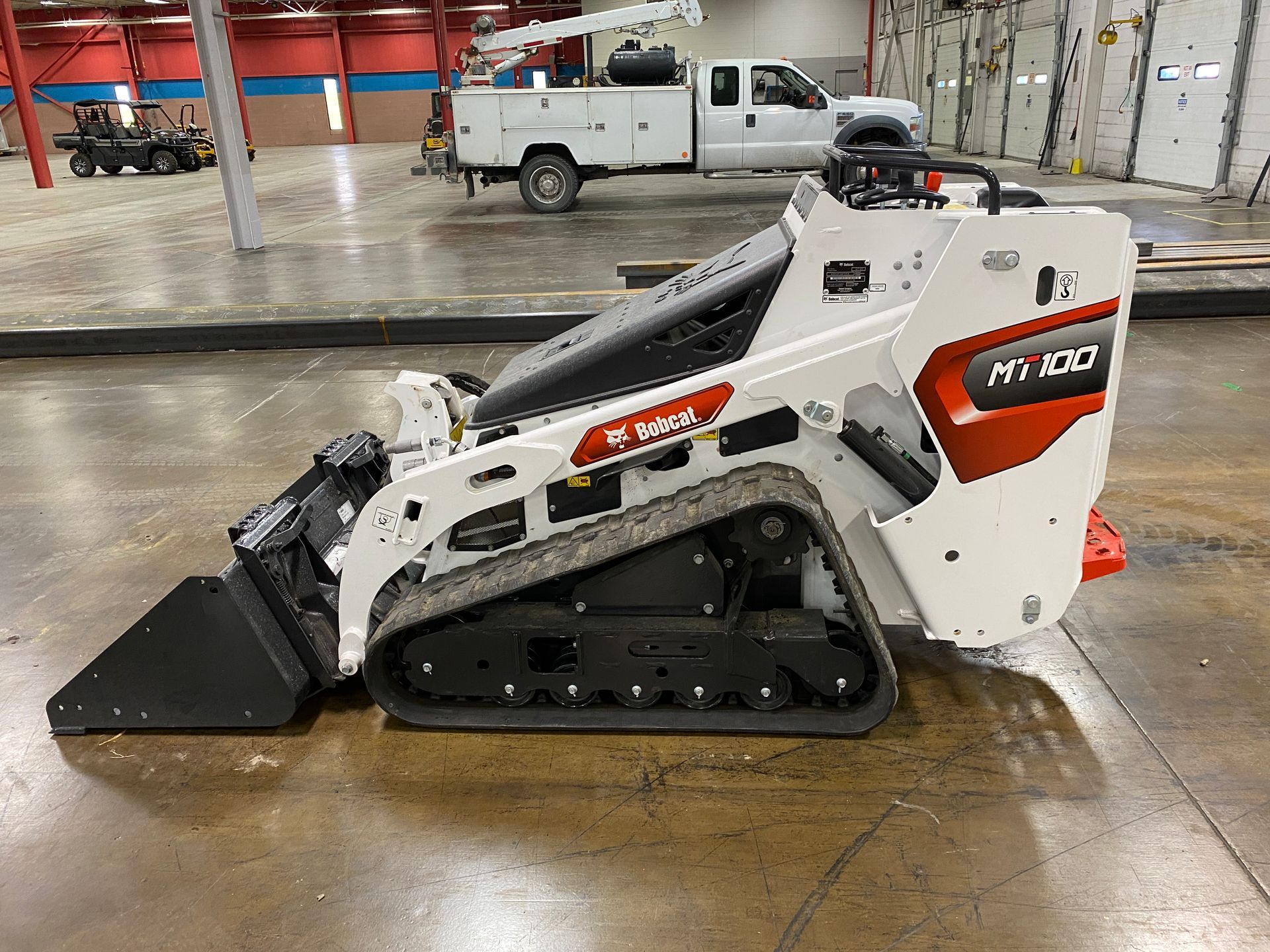 White Bobcat MT100 compact track loader with bucket in a warehouse, with a truck in the background.