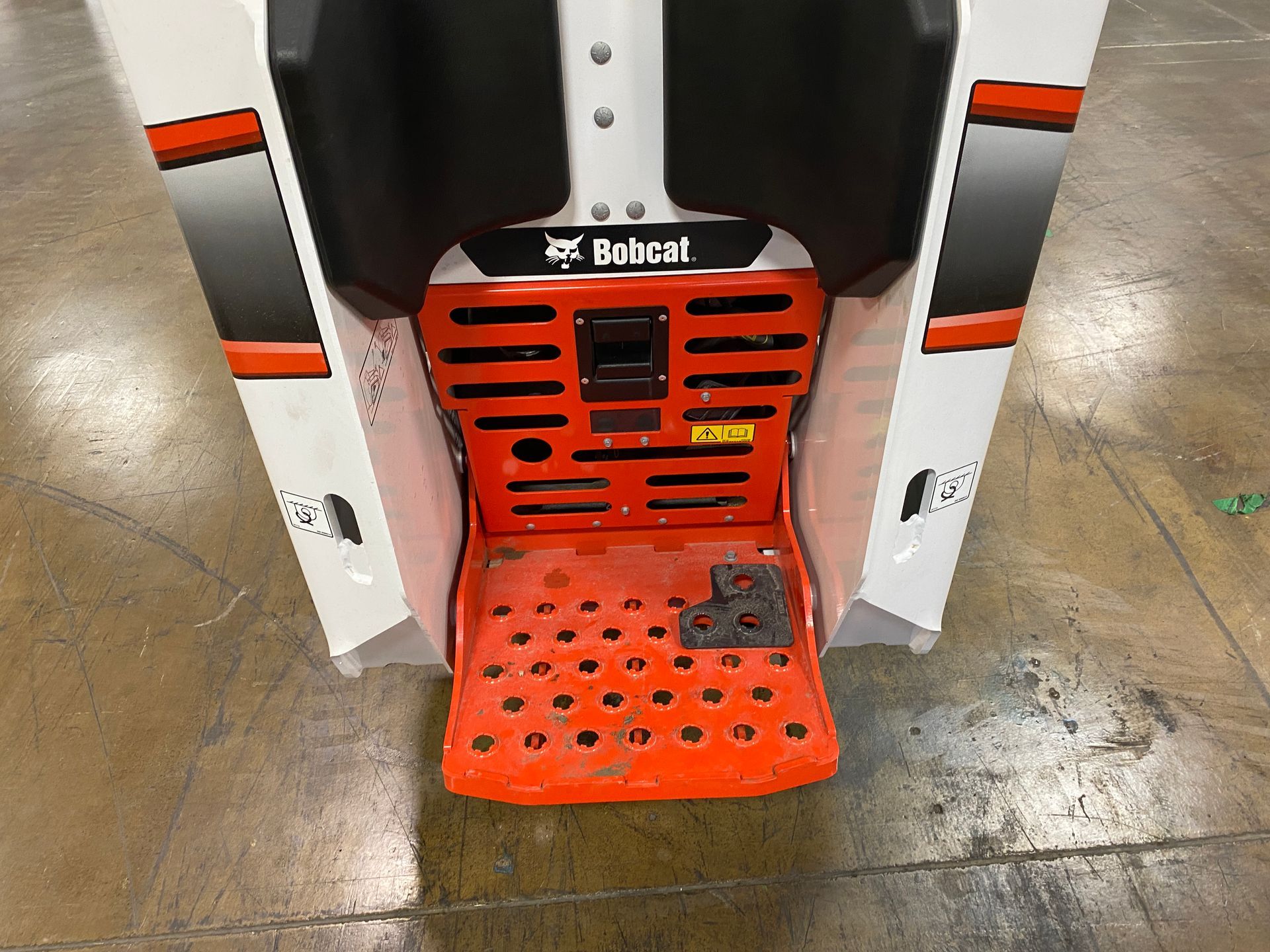 Rear view of an orange and white Bobcat machine with a footrest and a black seat.
