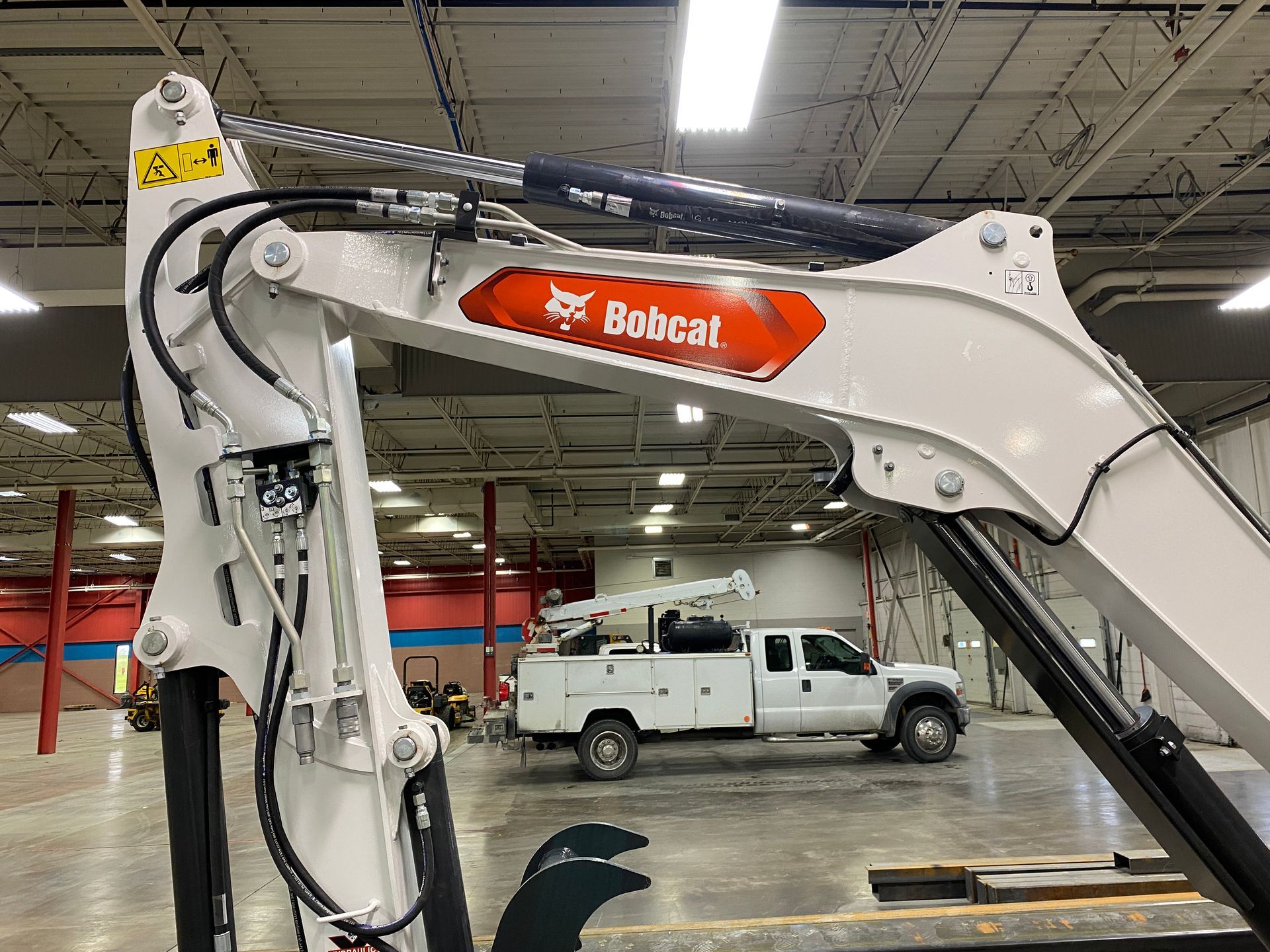 White Bobcat excavator arm with logo, with a white truck in the background. Inside a large building.