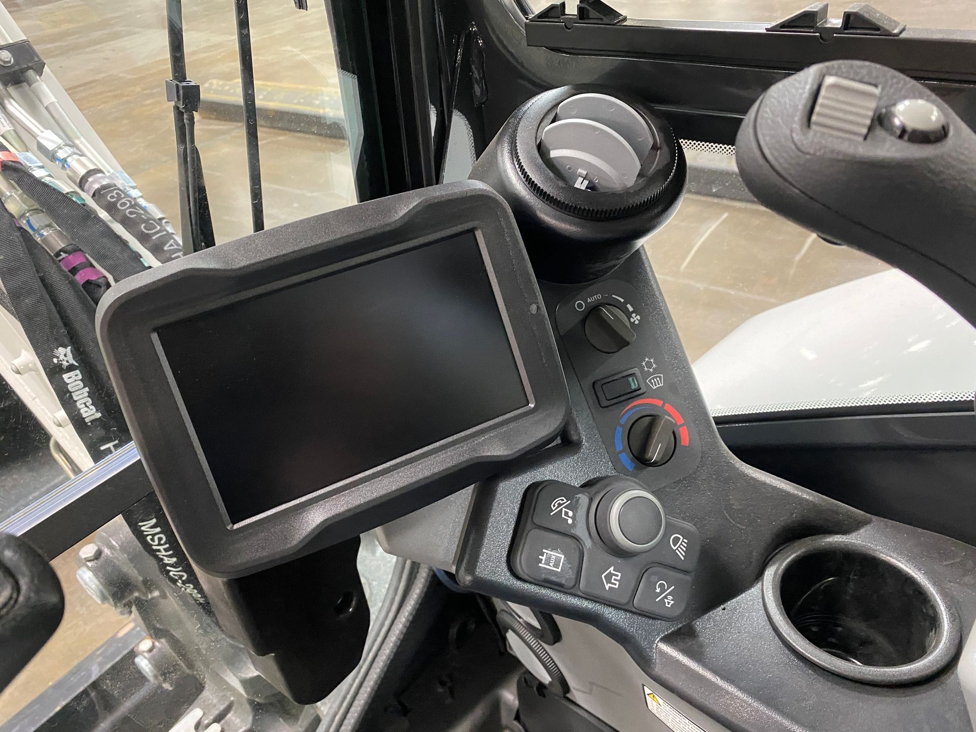Dashboard of a Bobcat heavy machine, including a screen, joystick, air vents, and cup holder.