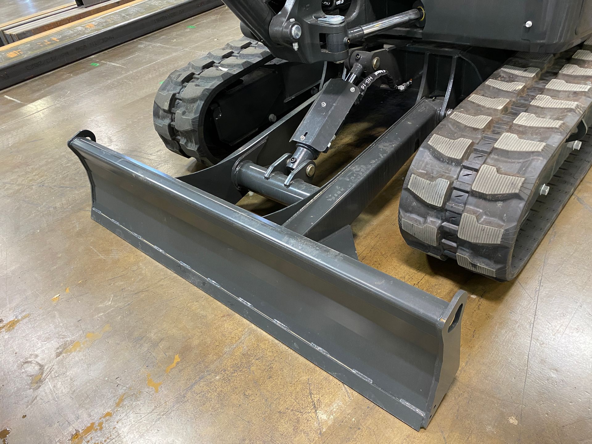 Mini excavator with a gray blade and black tracks on a concrete floor.